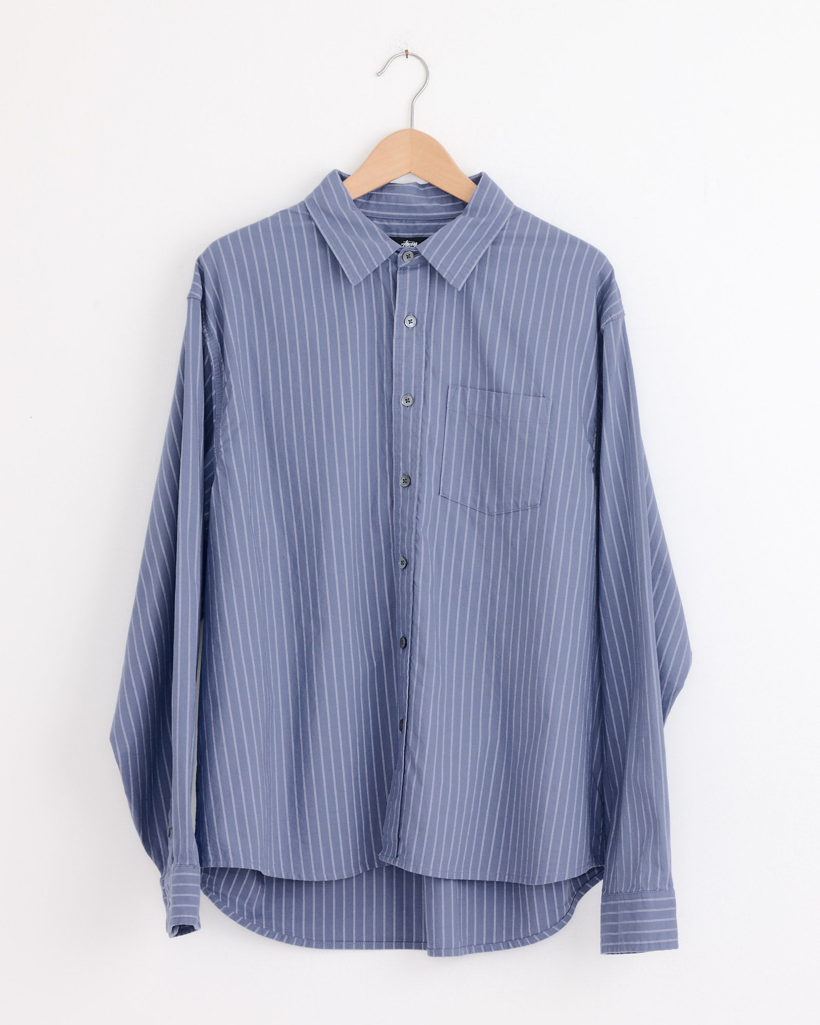 The Stussy Oxford Shirt in Blue Stripe is a boxy-fit, long-sleeve shirt with white pinstripes, a chest pocket, and classic detailing. It hangs on a wooden hanger against a white background.