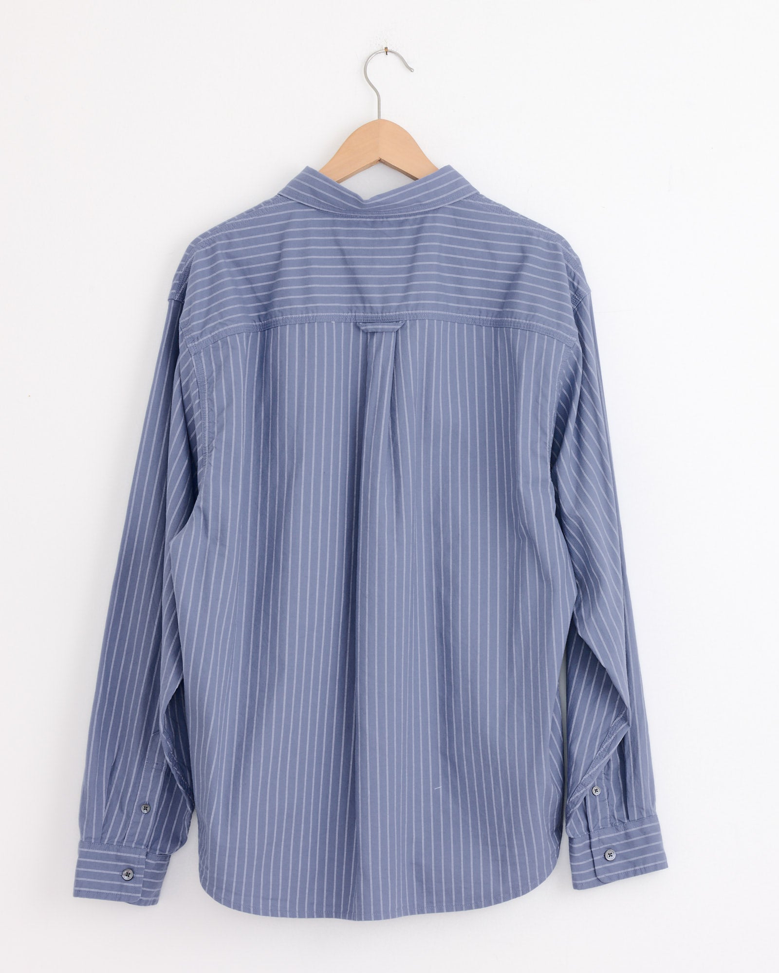 The Stussy Oxford Shirt in Blue Stripe, featuring white vertical stripes and a boxy fit, is displayed from the back on a wooden hanger against a white wall.