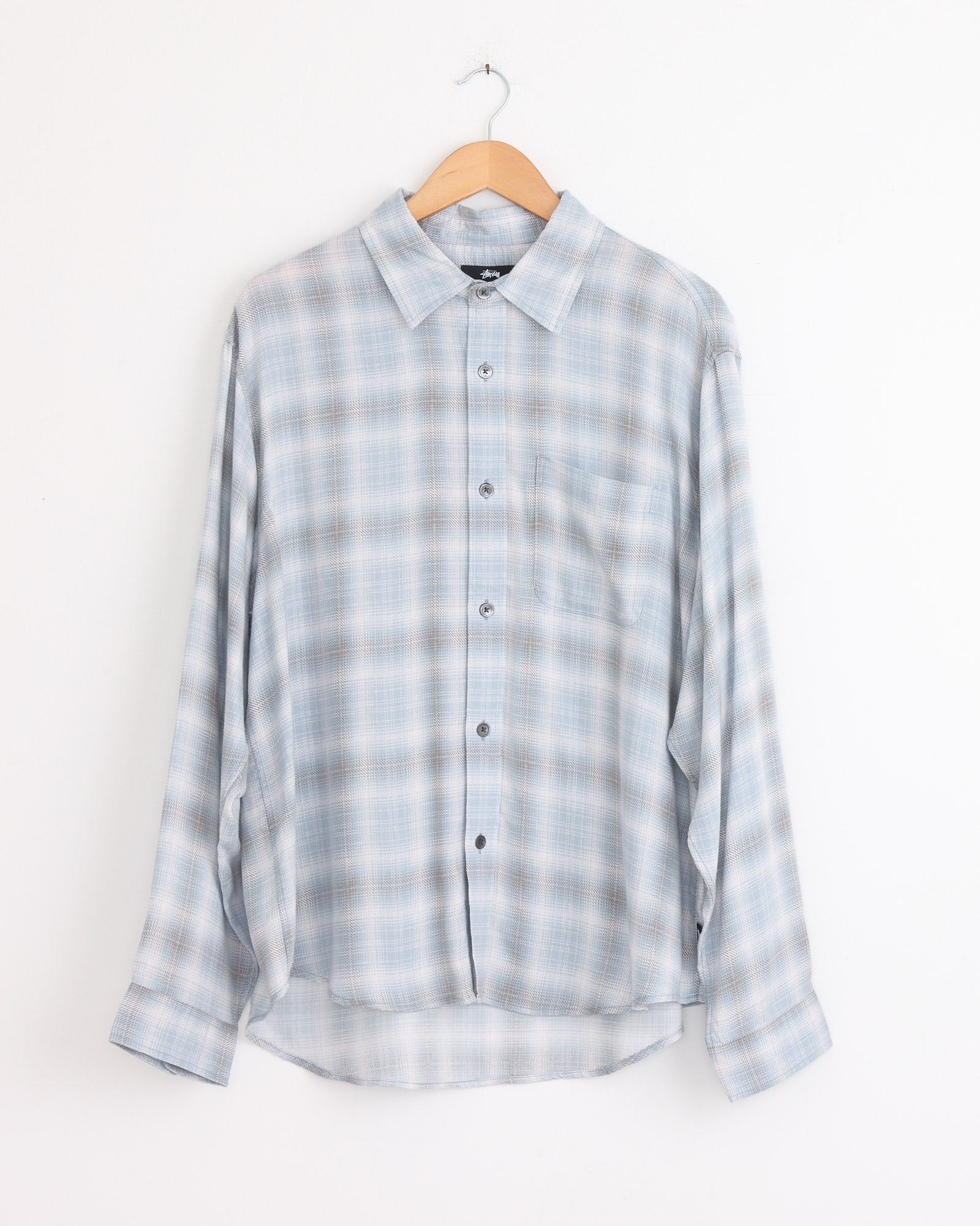 The Stussy Matthew Plaid Shirt in Light Blue, a brushed cotton flannel long-sleeve with gray accents, features a single chest pocket and curved hem. It hangs on a wooden hanger against a plain white wall.