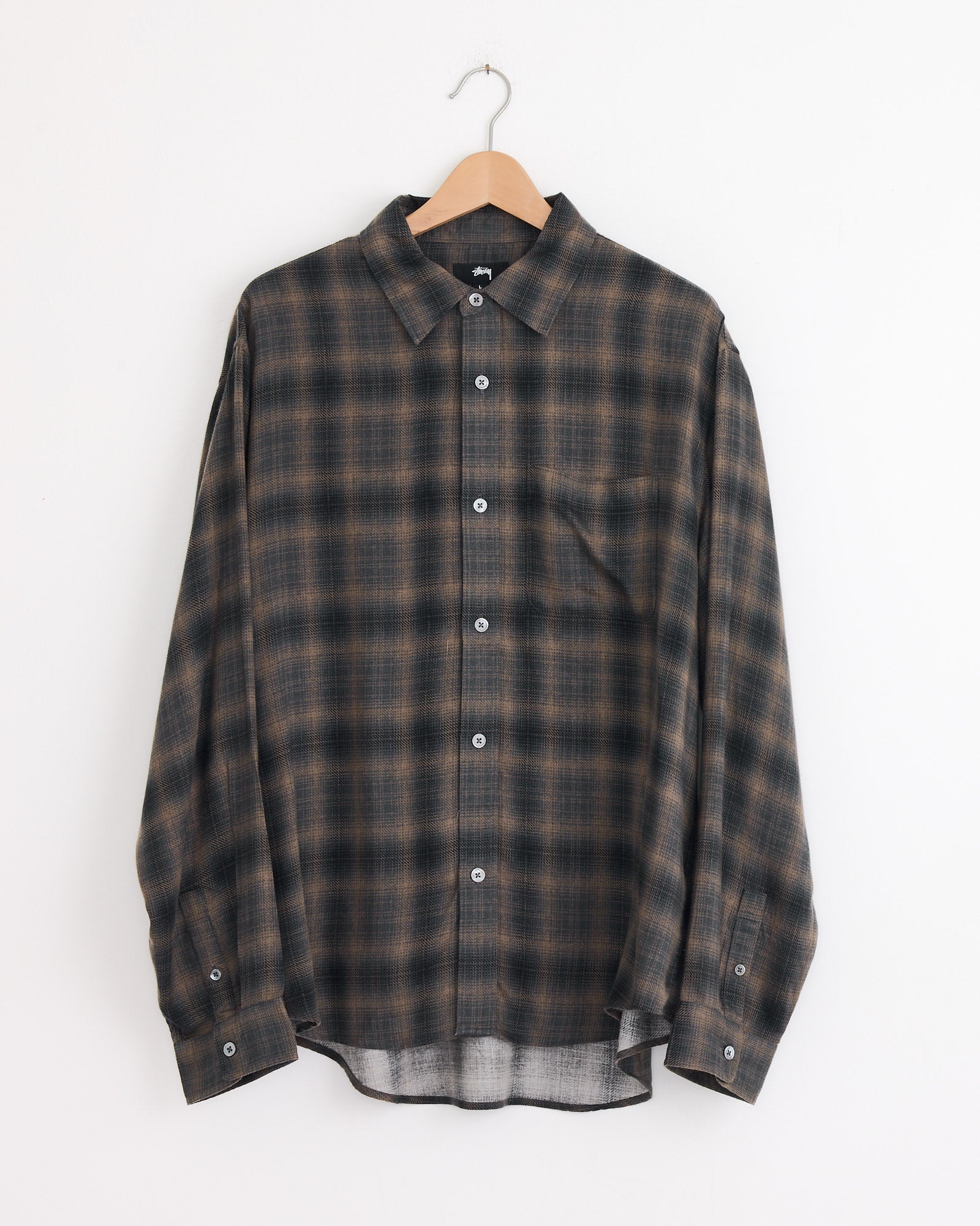 The Stussy Matthew Plaid Shirt in Brown, a classic-fit long-sleeve shirt with a brown and black pattern, crafted from soft brushed cotton flannel, hangs on a wooden hanger against a plain white wall.