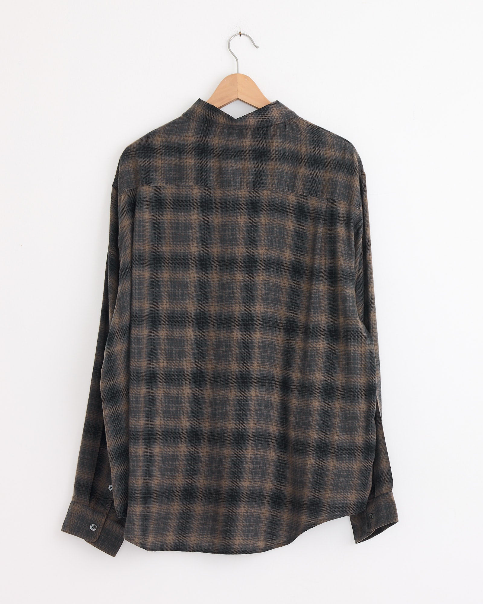 The Stussy Matthew Plaid Shirt in Brown, a classic-fit, long-sleeved button-up with a brown and gray plaid pattern, is displayed from the back on a wooden hanger against a plain white wall. Made from brushed cotton flannel.