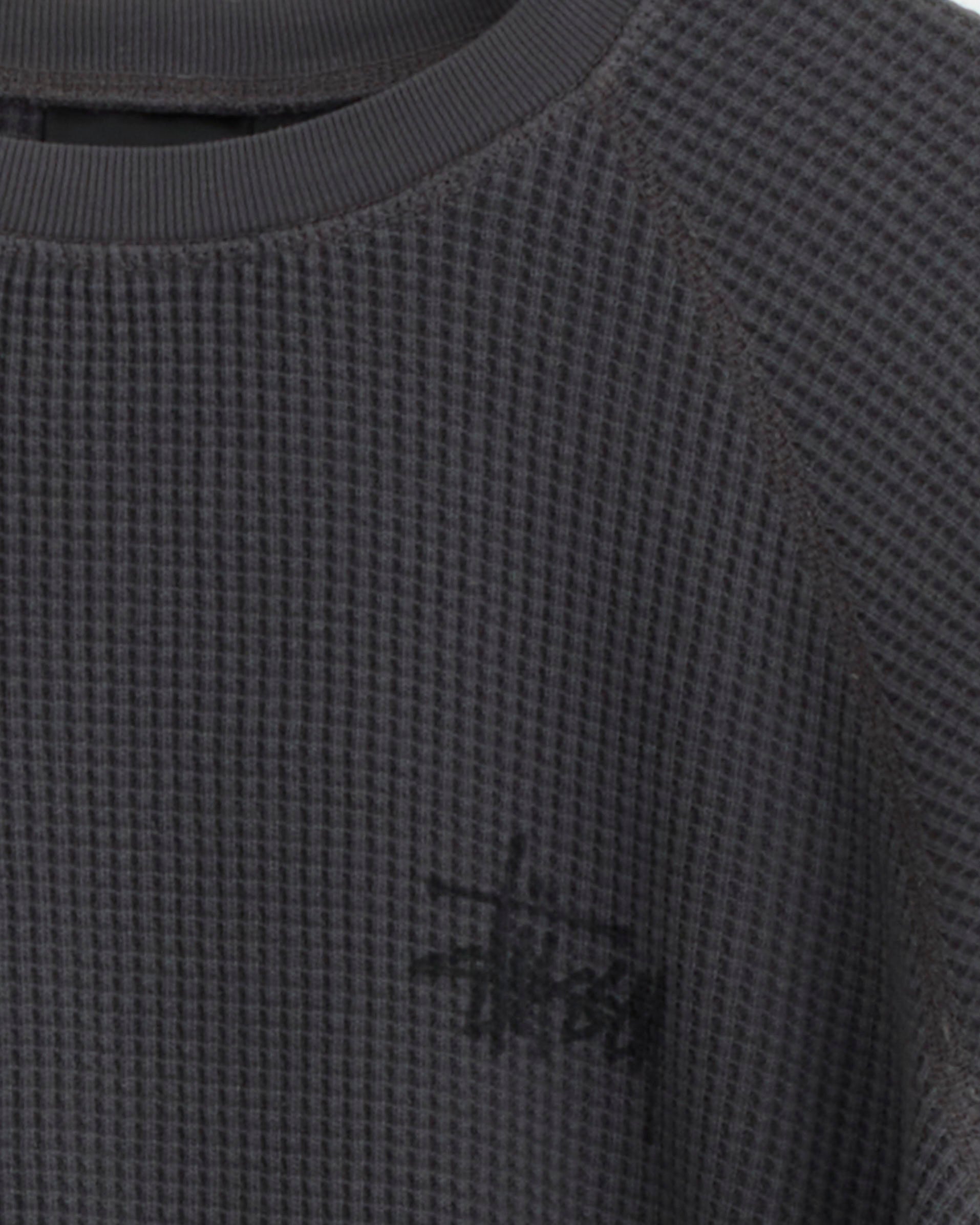 Close-up of a washed black, waffle-knit cotton sweater with ribbed collar. The Stussy logo appears on the bottom right, highlighting this staple as the Stussy Raglan Thermal Basic Stock.