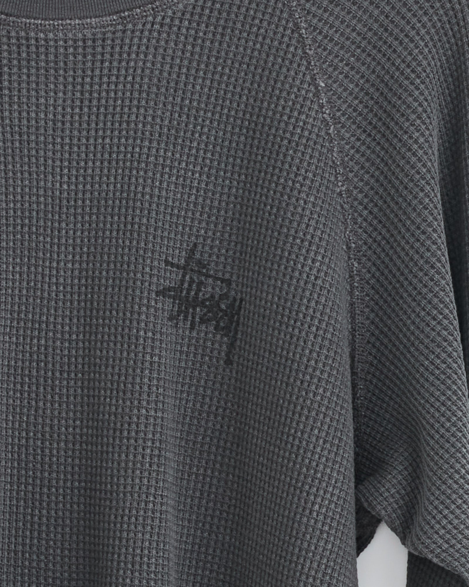 A close-up of the Stussy Raglan Thermal Basic Stock shirt in washed black, featuring a waffle-knit cotton design and the Stussy logo printed in black on the upper left chest.