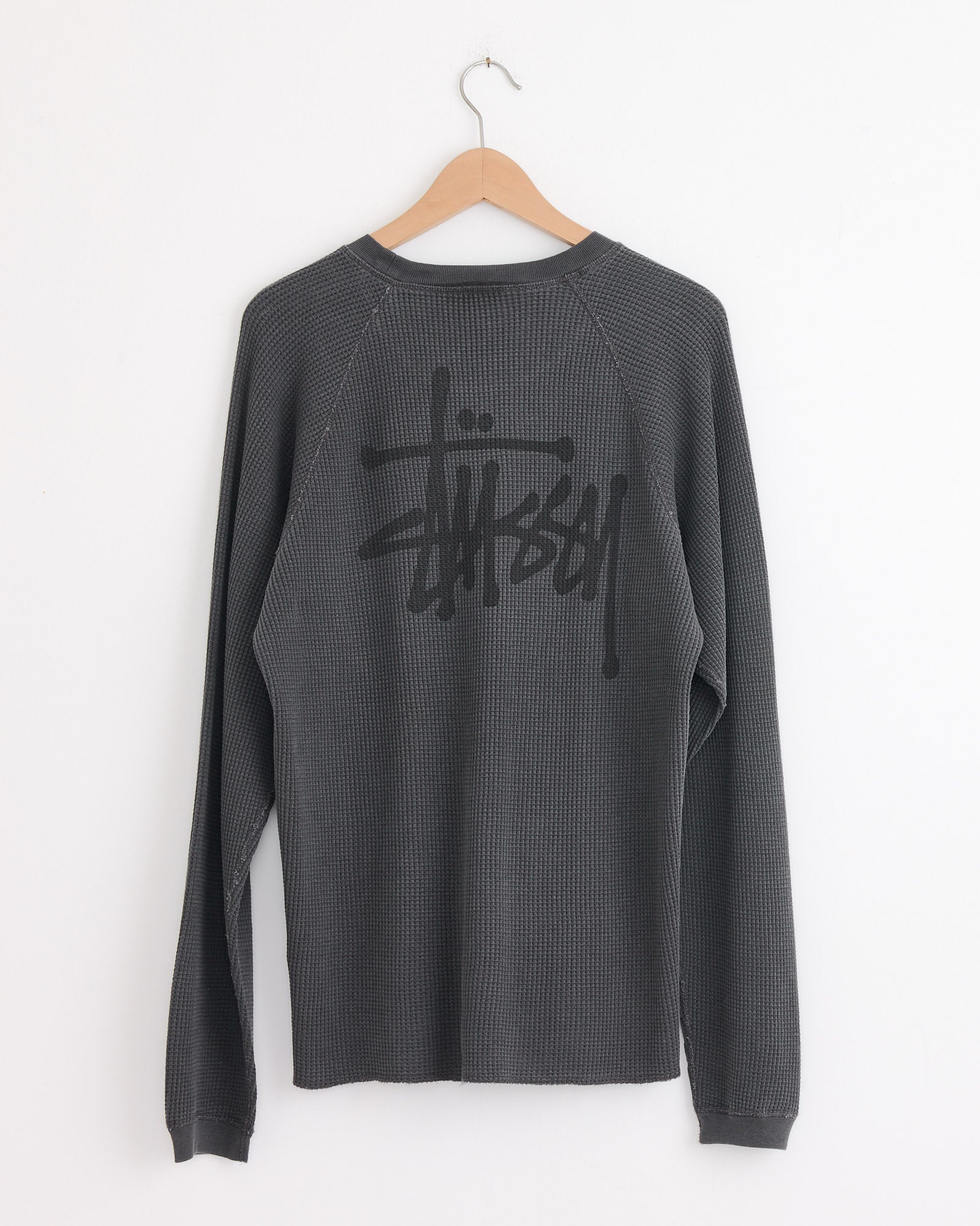 A Raglan Thermal Basic Stock in Washed Black by Stussy hangs on a wooden hanger against a white wall. Made from waffle-knit cotton, its back displays a large black Stussy logo in graffiti-style text.