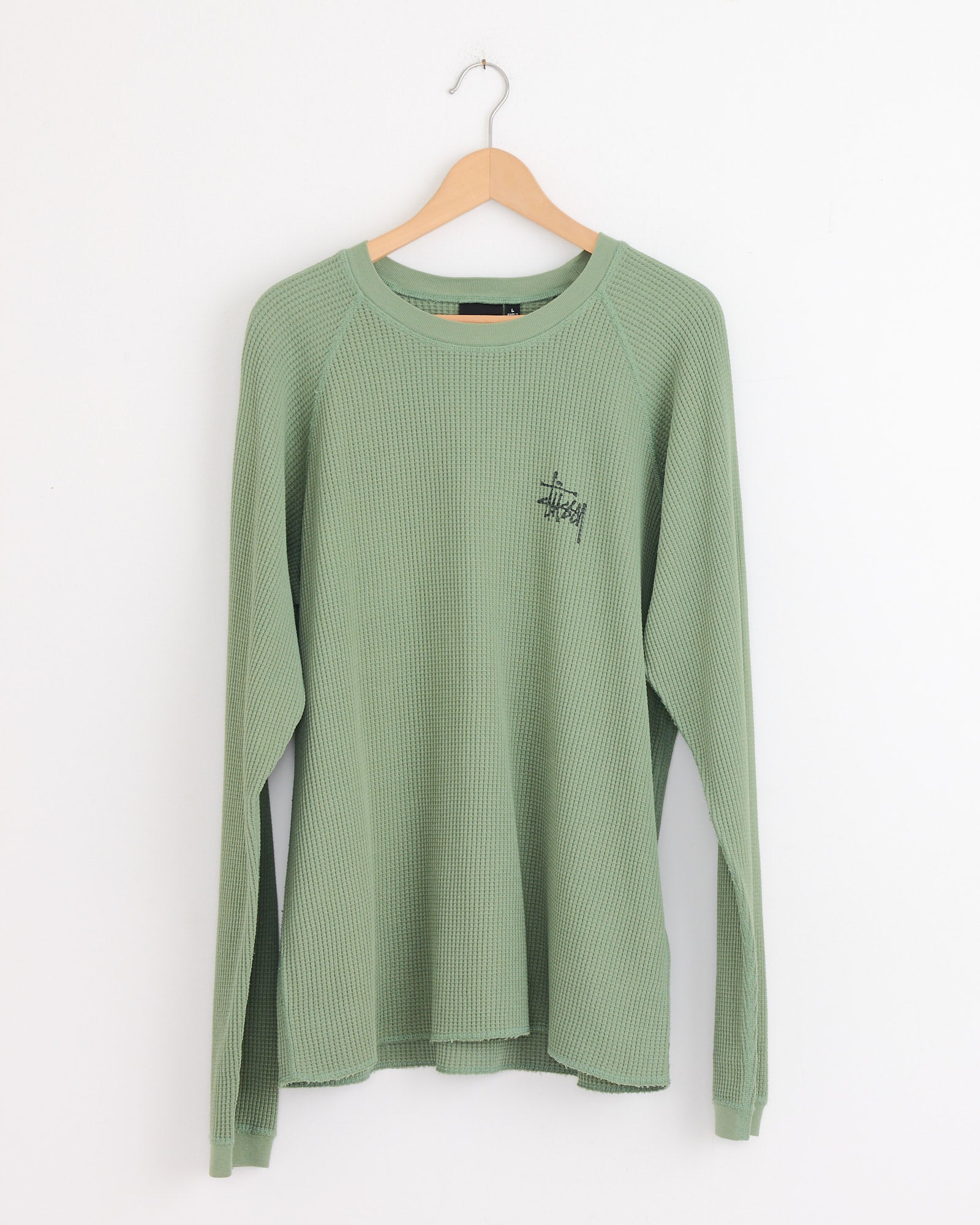 The Stussy Raglan Thermal Basic Stock in Sage is a long-sleeve, waffle-knit cotton shirt with a round neckline and small black text on the left chest, shown hanging on a wooden hanger against a white wall.