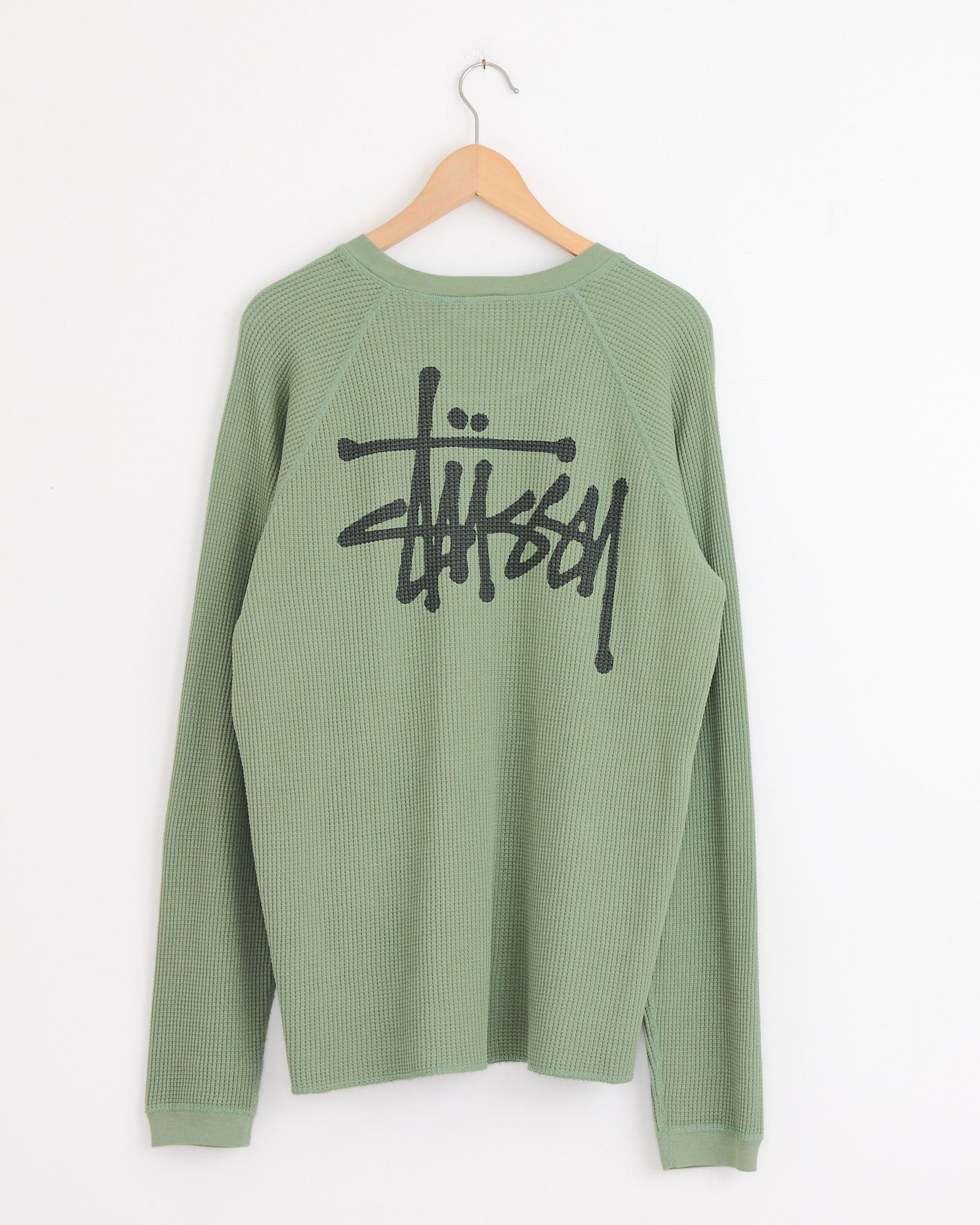 The Stussy Raglan Thermal Basic Stock in Sage is a light green, long-sleeve, waffle-knit cotton shirt with a ribbed texture and features a large black Stussy graffiti-style logo on the back.