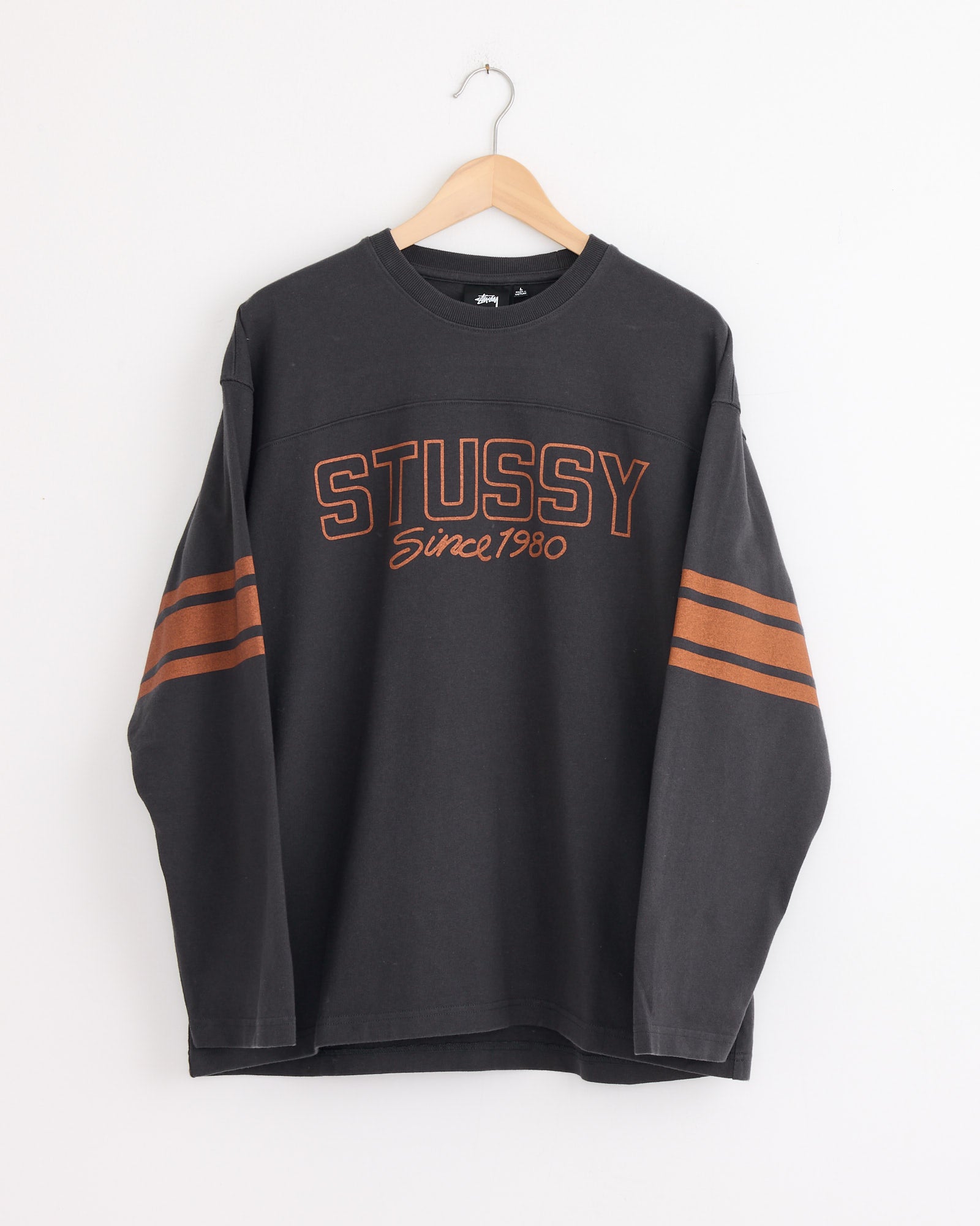 The Stussy Football Crew in Black is a heavyweight cotton long-sleeve sweatshirt with orange stripes and "STUSSY Since 1980" in orange on the front, shown hanging on a wooden hanger against a white wall.