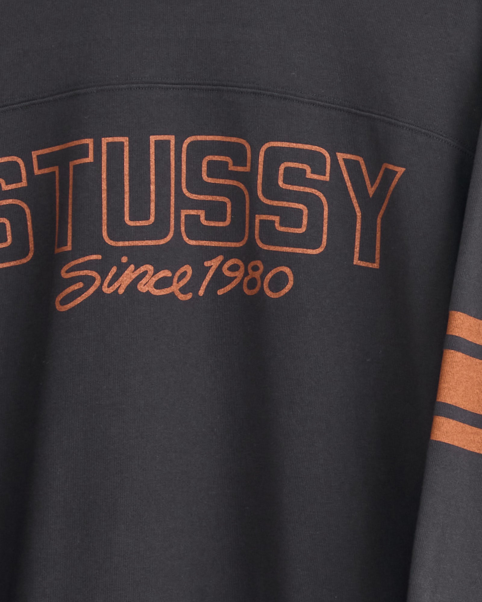 The Stussy Football Crew in Black is a heavyweight cotton long-sleeve shirt with bold orange "STUSSY," "Since 1980" in cursive below, and two orange sleeve stripes.