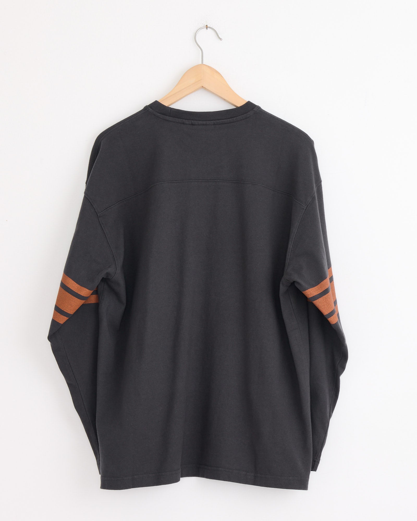 A Stussy Football Crew in Black hangs on a wooden hanger against a white wall, showing two orange stripes near the elbows. The unisex heavyweight cotton long-sleeve tee is displayed with the back facing forward.