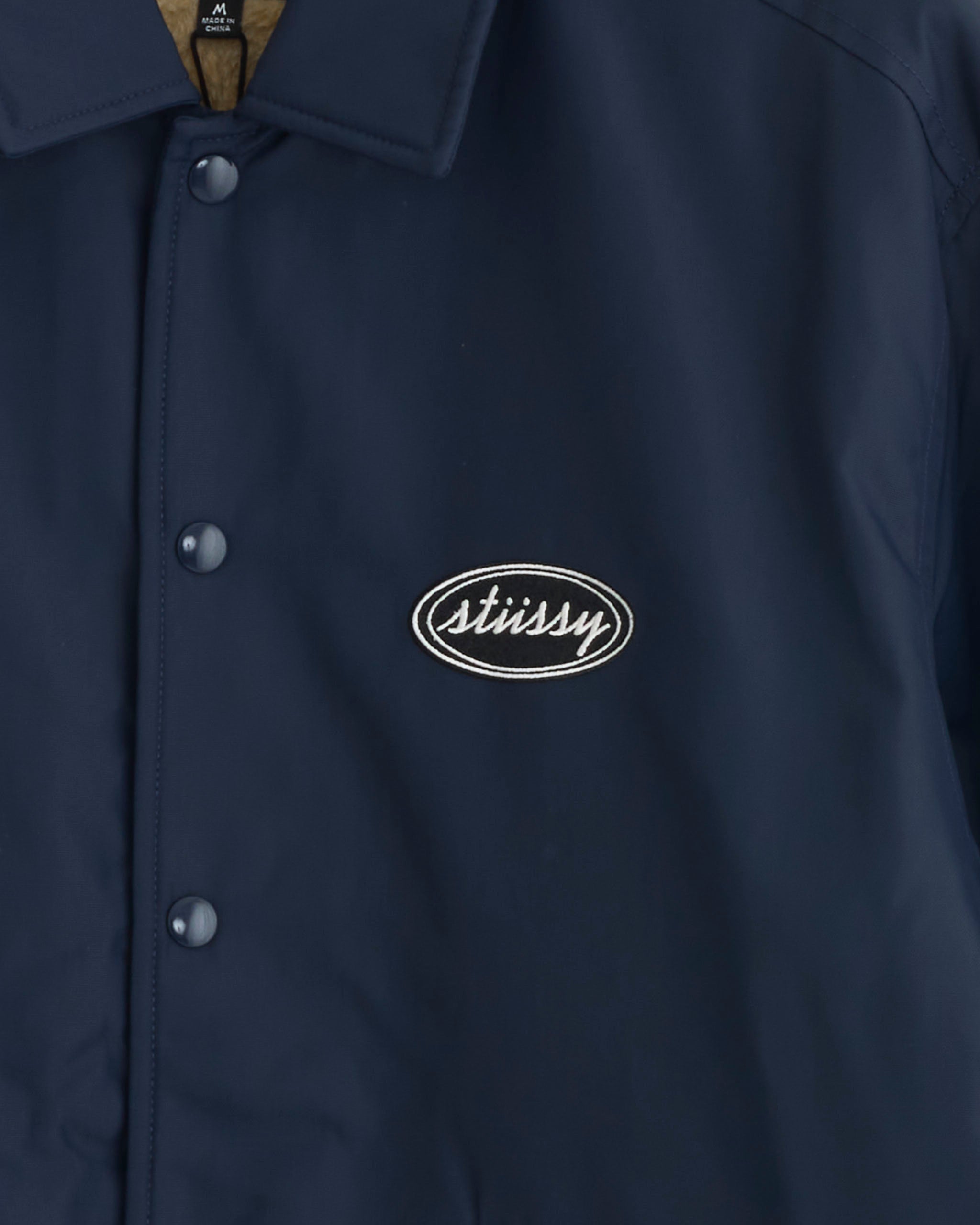 Close-up of the Stussy Coaches Jacket in Navy, featuring snap buttons and a white oval Stussy logo patch embroidered on the left chest, highlighting its classic style.