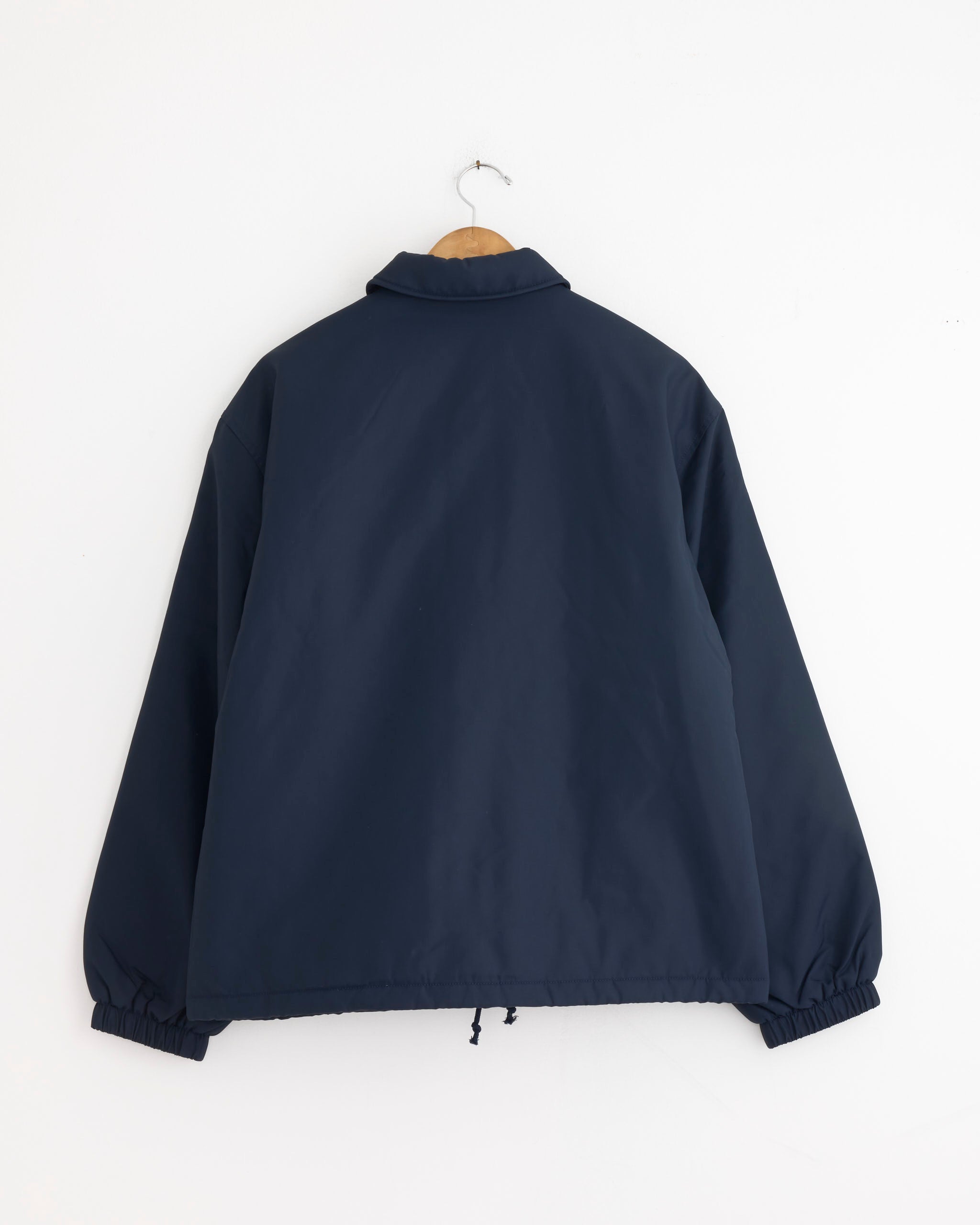 The Stussy Coaches Jacket in Navy, featuring a collar and elastic cuffs, hangs on a wooden hanger against a white wall. The nylon oxford jacket is displayed from the back.