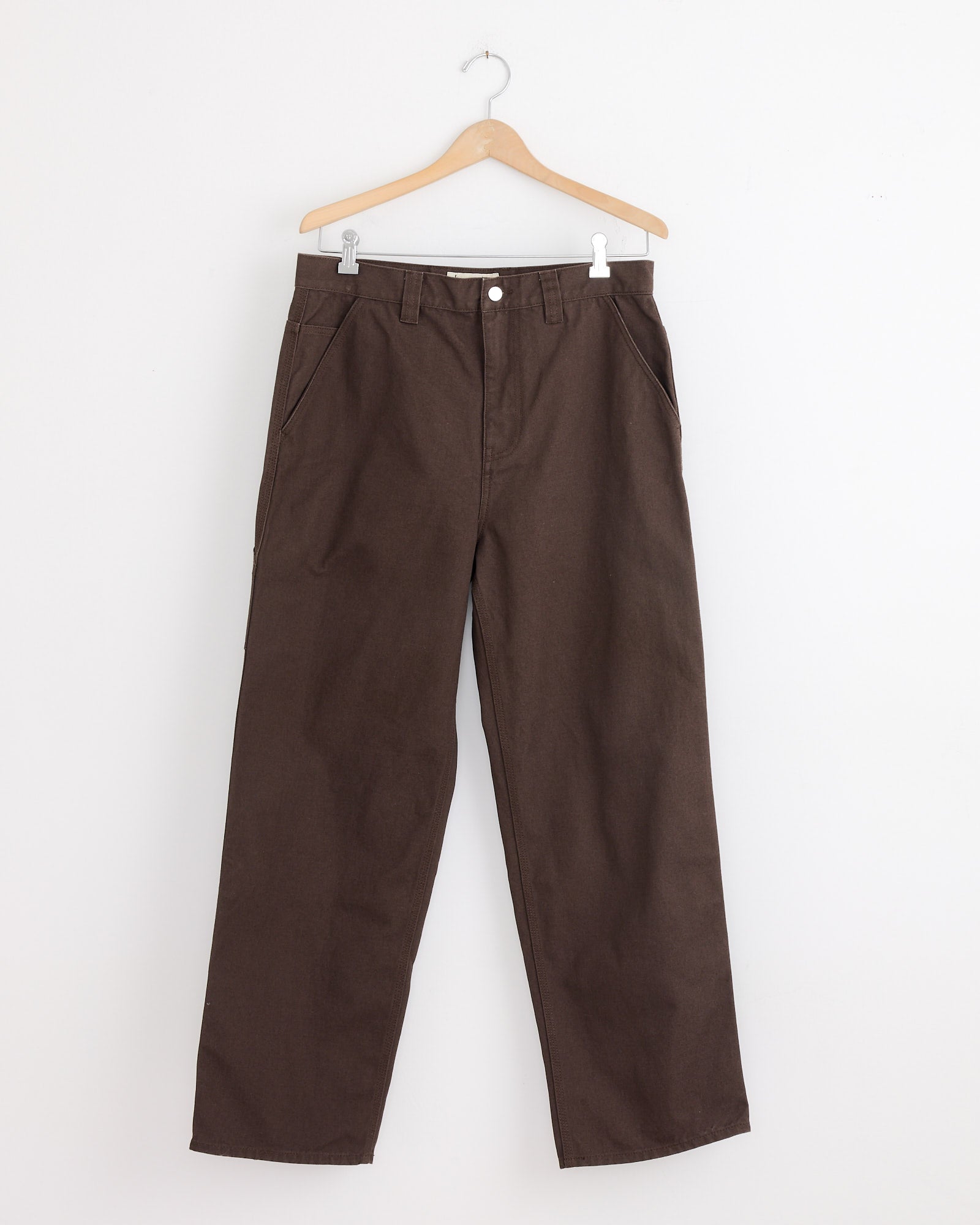 The Stussy Carpenter Pant in Brown hangs on a wooden hanger against a plain white wall, ideal for stylish workwear.