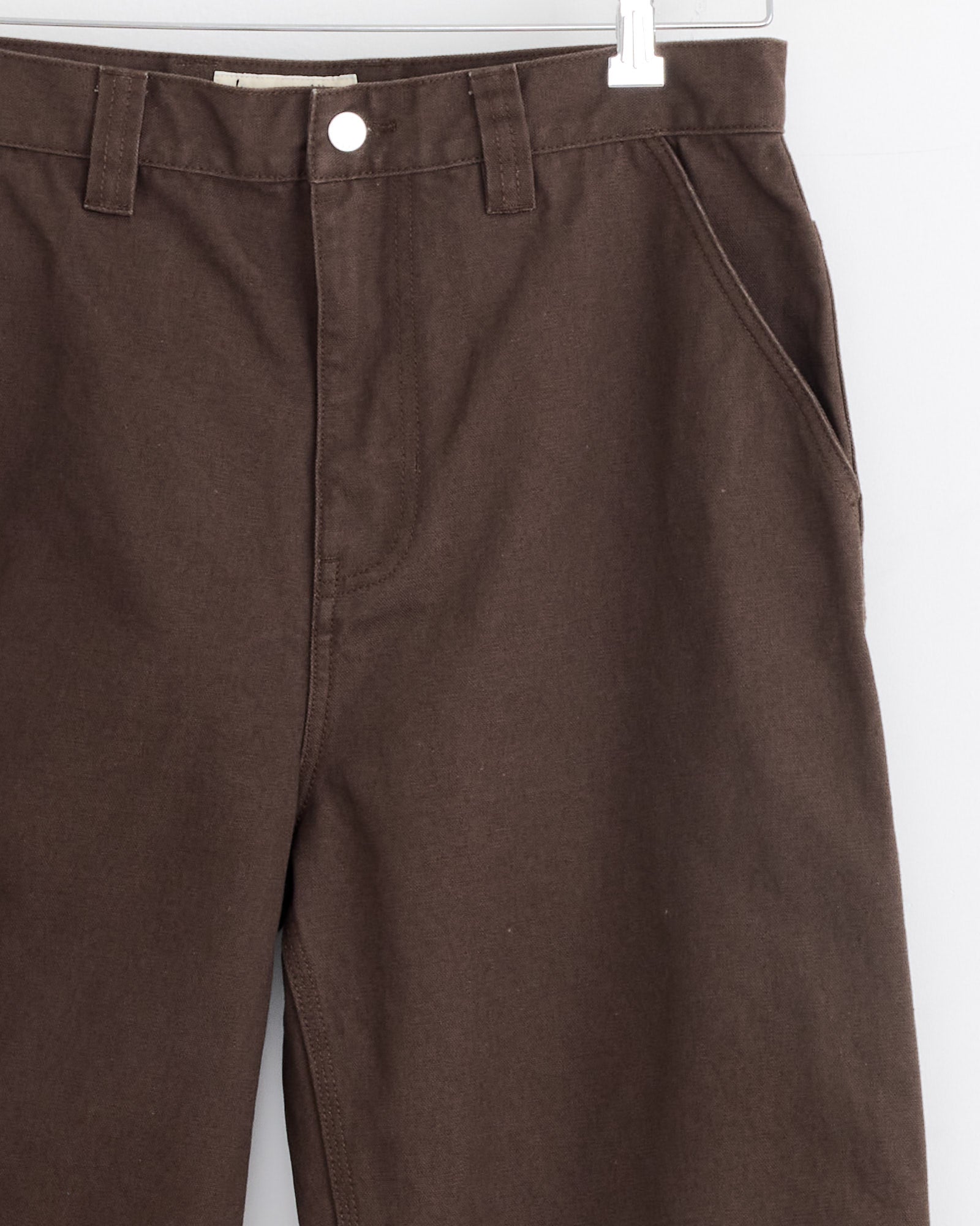 Close-up of the Stussy Carpenter Pant in Brown, displayed on a hanger with focus on waistband, button, zipper, belt loops, and front pockets against a clean white background.