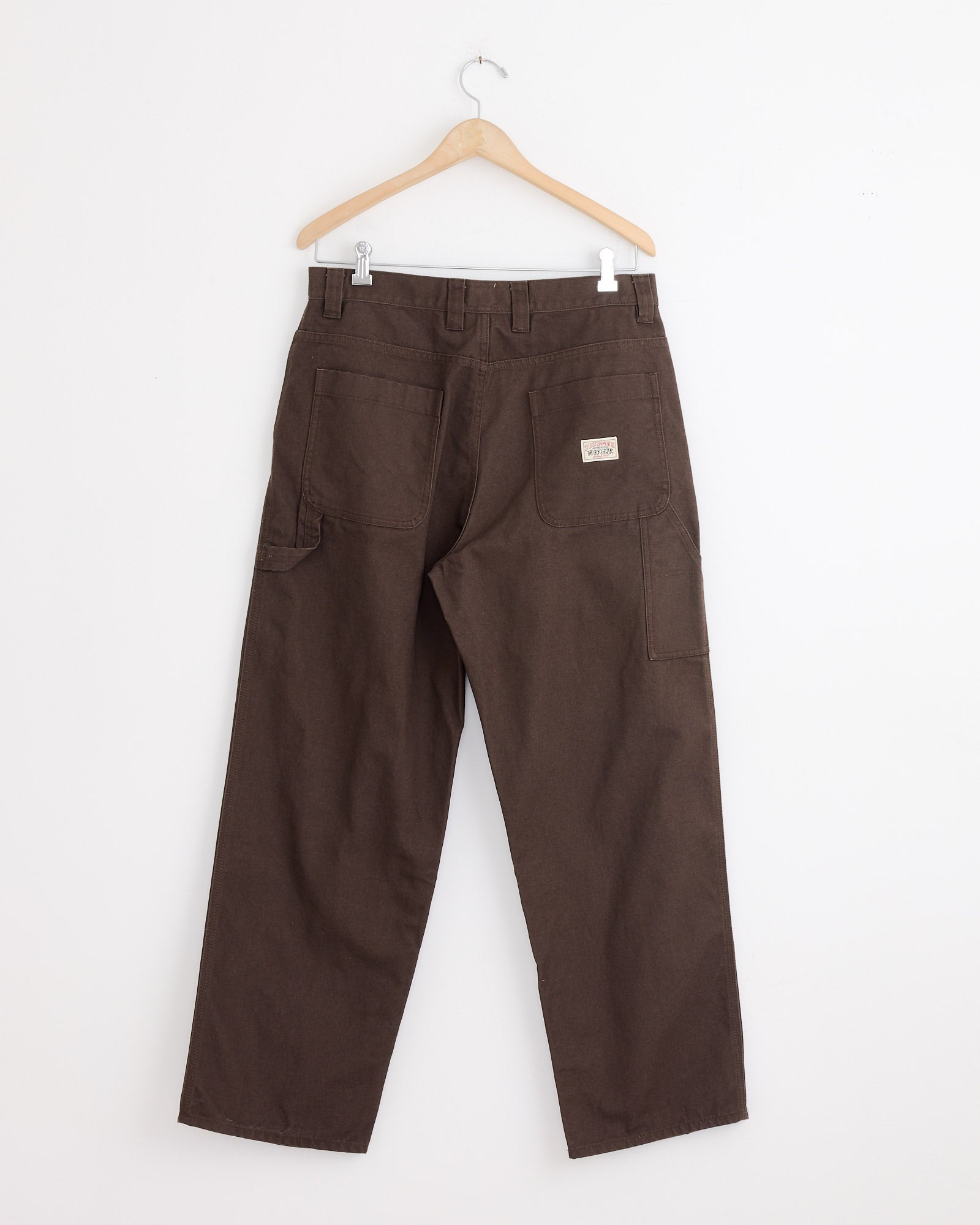 The Stussy Carpenter Pant in Brown hangs from a wooden hanger against a white wall, shown from the back with large workwear-style back pockets and a small rectangular patch above the right pocket.