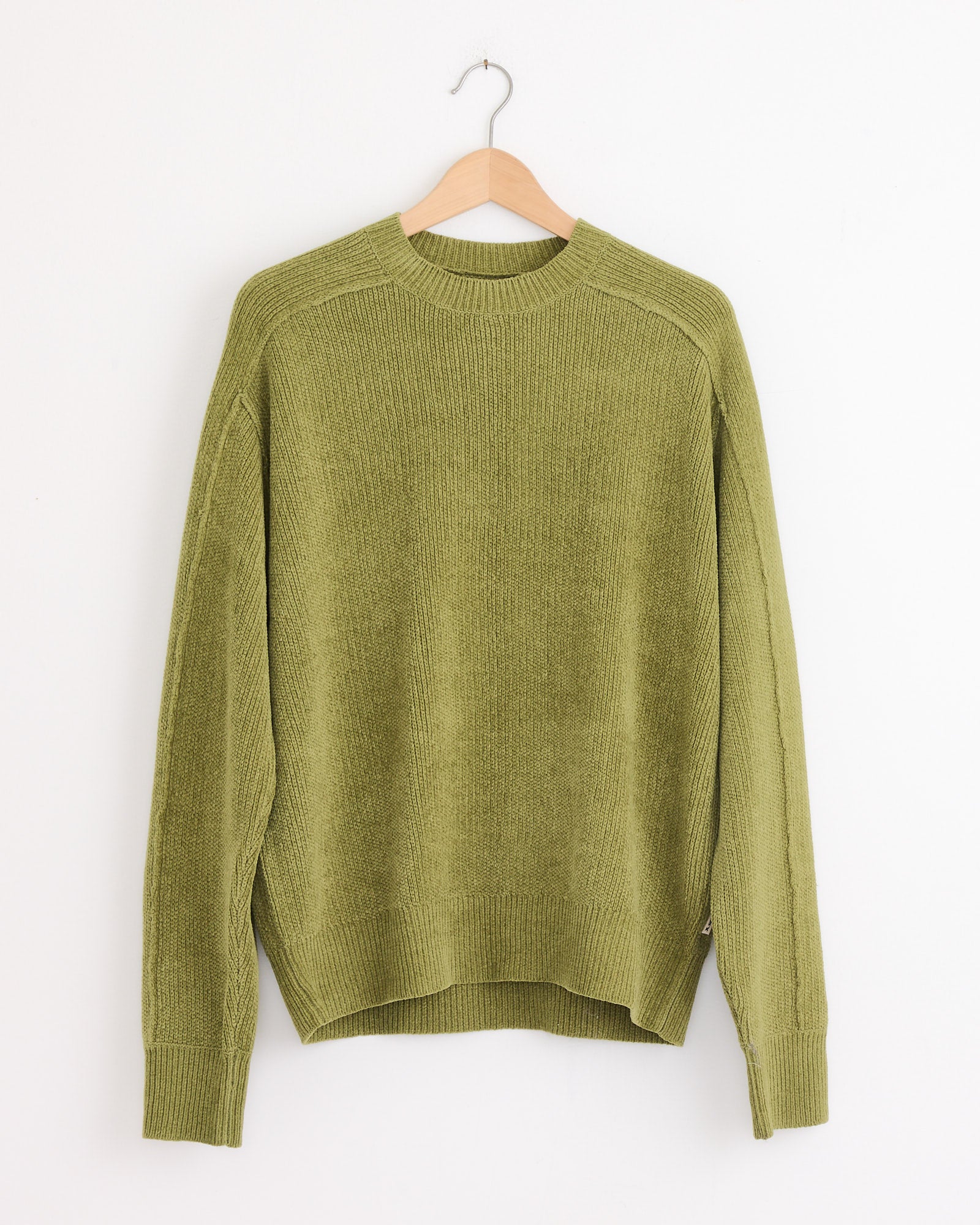 The Stussy Exposed Seam Sweater in Green, made from soft cotton chenille with long sleeves, hangs on a wooden hanger against a plain white wall.