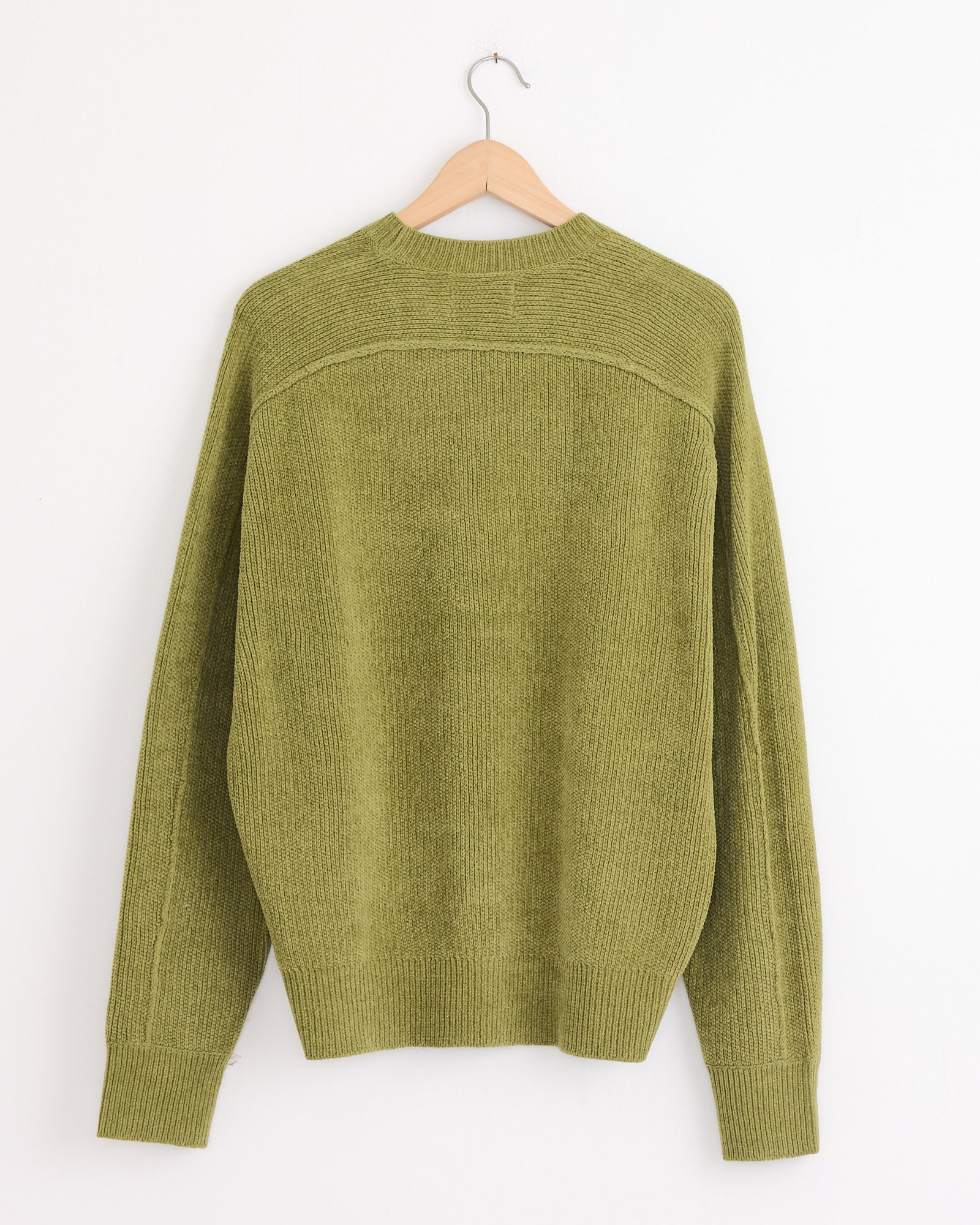 The Stussy Exposed Seam Sweater in Green, crafted from soft cotton chenille, hangs on a wooden hanger against a plain white wall and is displayed from the back.