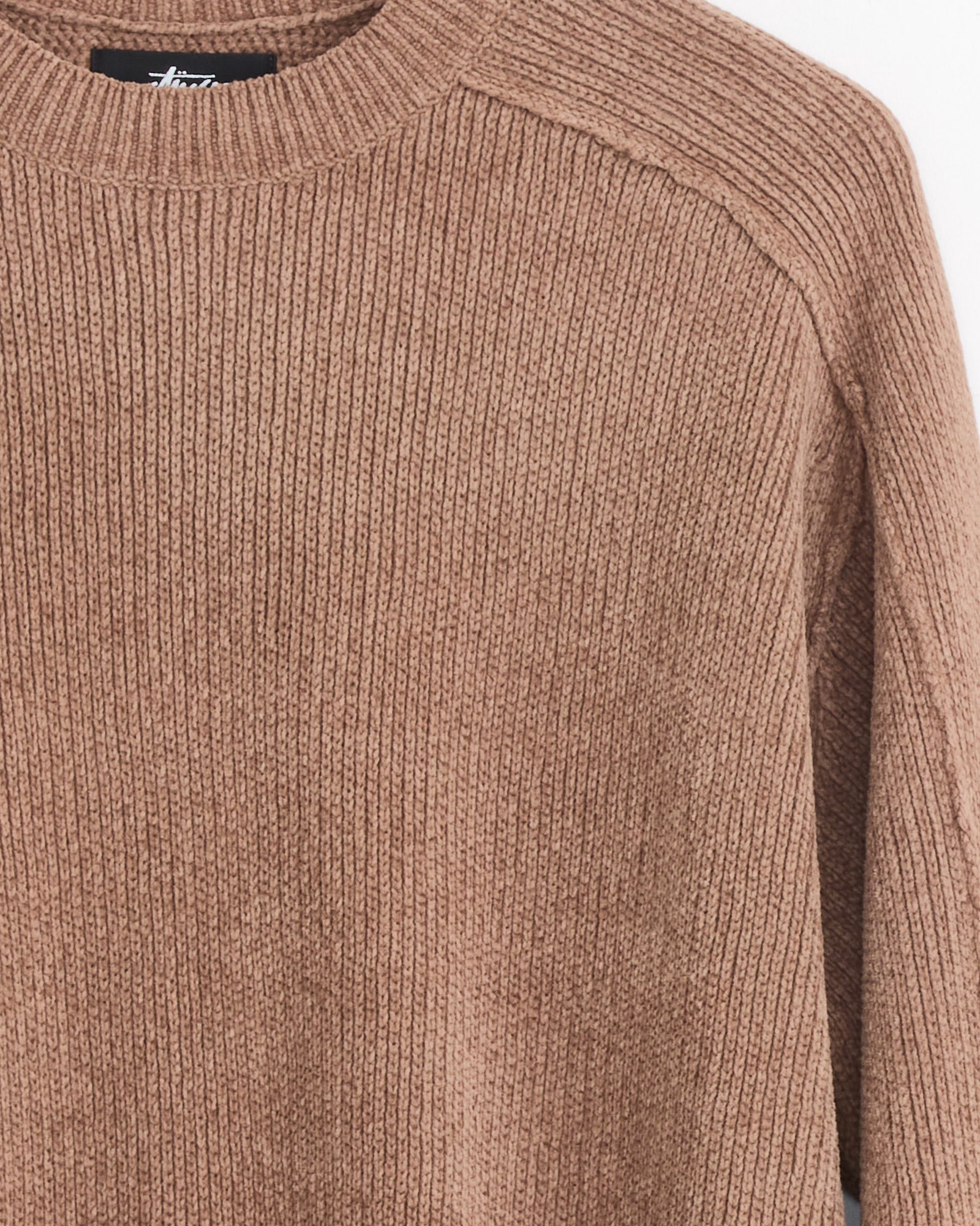 A close-up of the Stussy Exposed Seam Sweater in Brown highlights its ribbed knit texture, exposed seam stitching, round neckline, and visible sleeve detail.