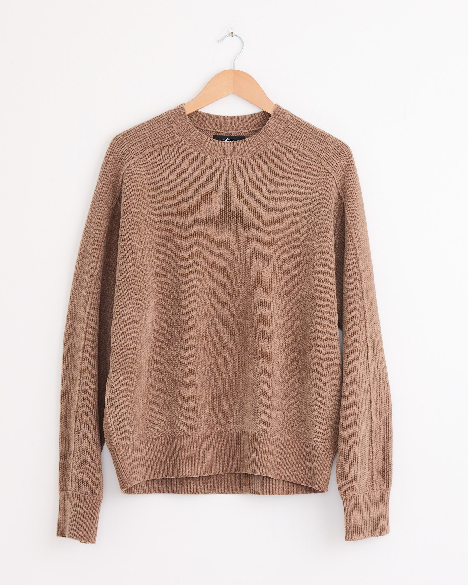 The Stussy Exposed Seam Sweater in Brown, featuring ribbed long sleeves and soft cotton chenille, hangs on a wooden hanger against a plain white wall.
