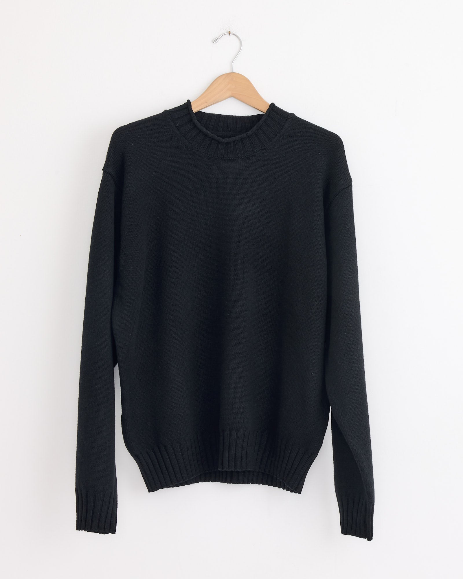 The Stussy Tonal Mock Neck Sweater in Black, featuring ribbed cuffs, hem, and neckline, hangs on a wooden hanger against a white wall.
