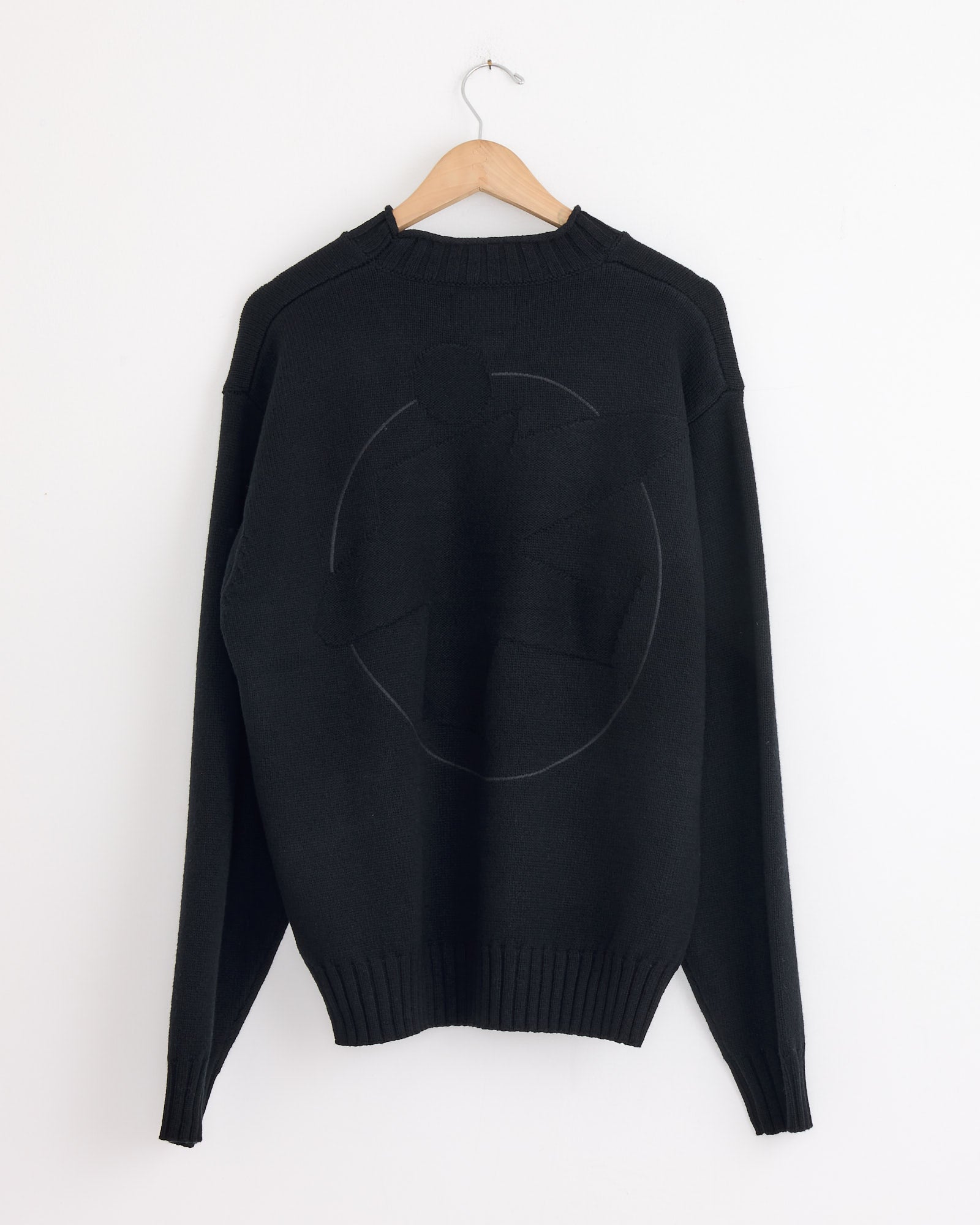 A black Stussy Tonal Mock Neck Sweater hangs on a wooden hanger against a white wall, showcasing a subtle circular abstract design in a slightly different black shade on the back.