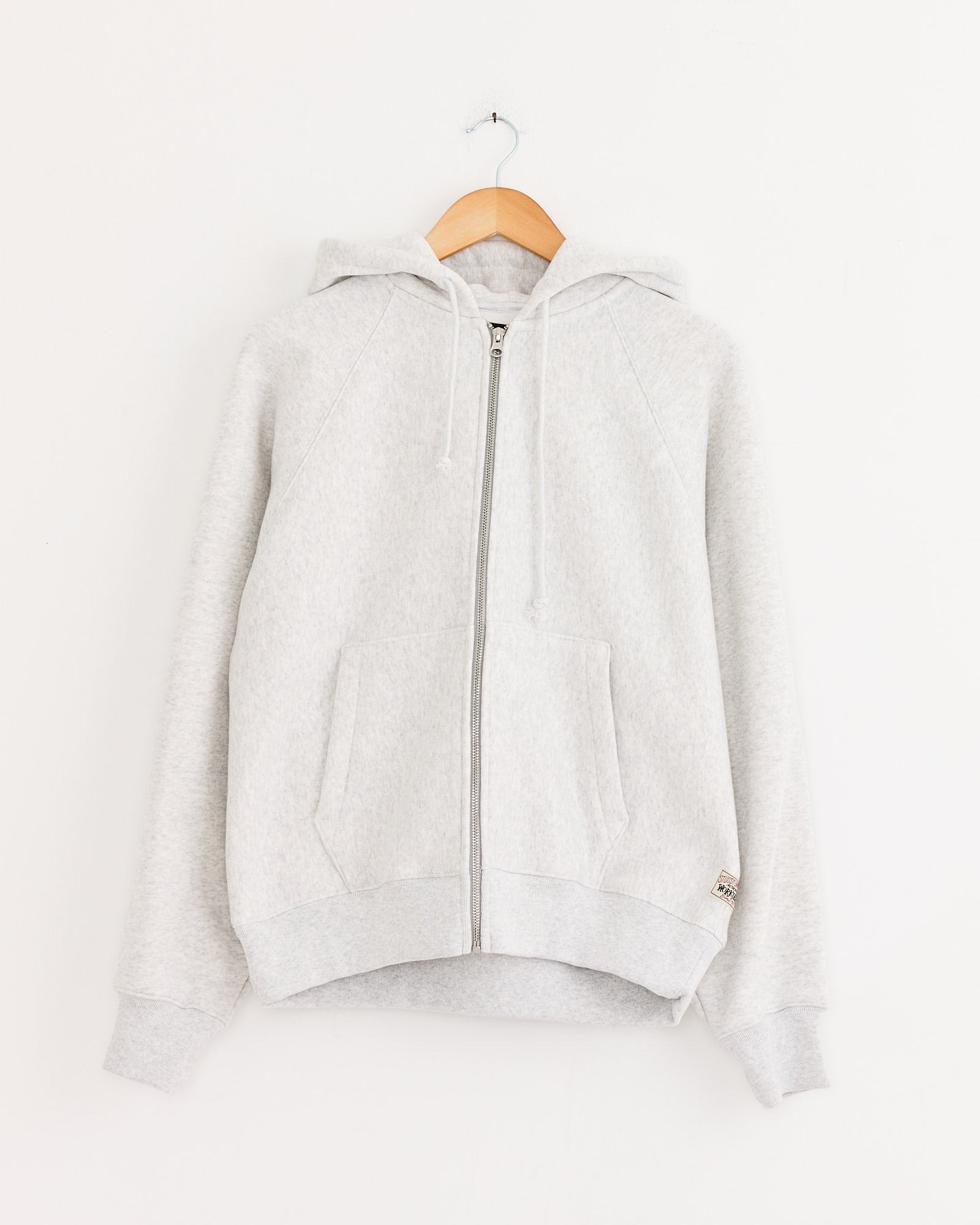 The Stussy Raglan Zip Hoodie in Ash Heather, crafted from heavyweight cotton with a hood and drawstrings, is displayed on a wooden hanger against a plain white wall.