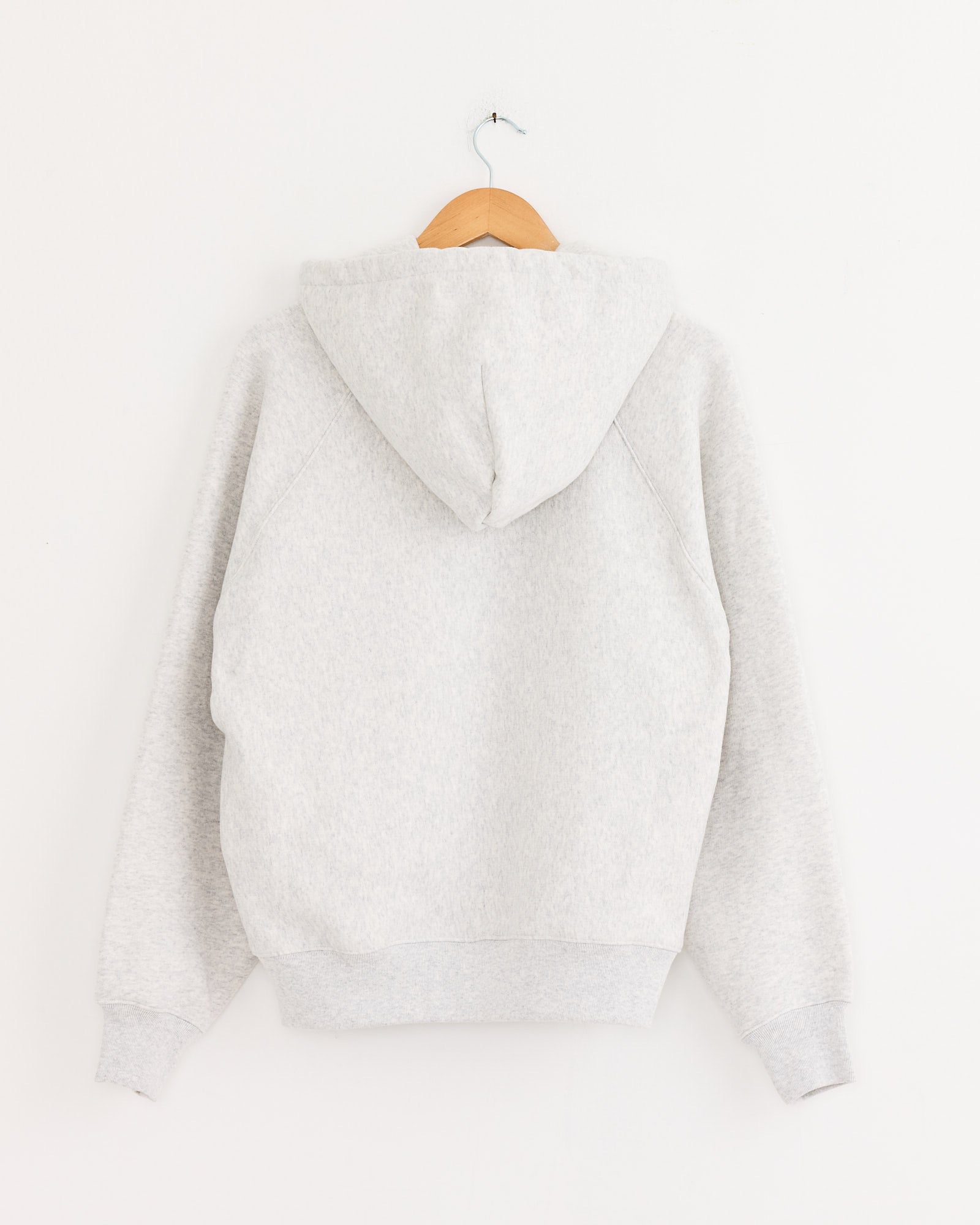 The Stussy Raglan Zip Hoodie in Ash Heather, crafted from heavyweight cotton, hangs on a wooden hanger against a plain white wall with the back facing forward.