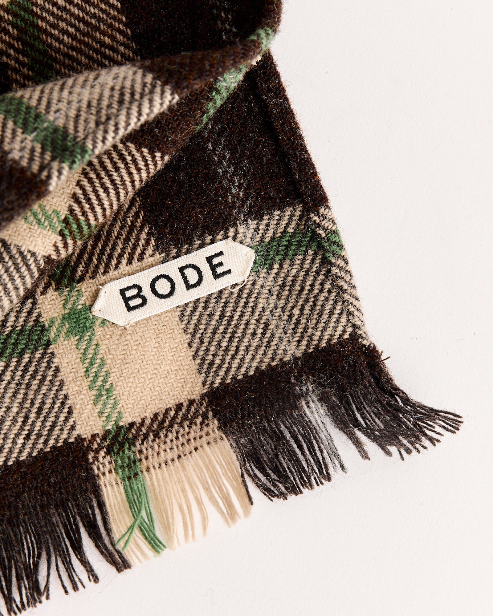 The Bode Woodcrest Scarf in Brown/Green features a plaid wool design with brown, beige, green, and black patterns, fringed edges, and a signature white BODE label, shown laid on a light surface.