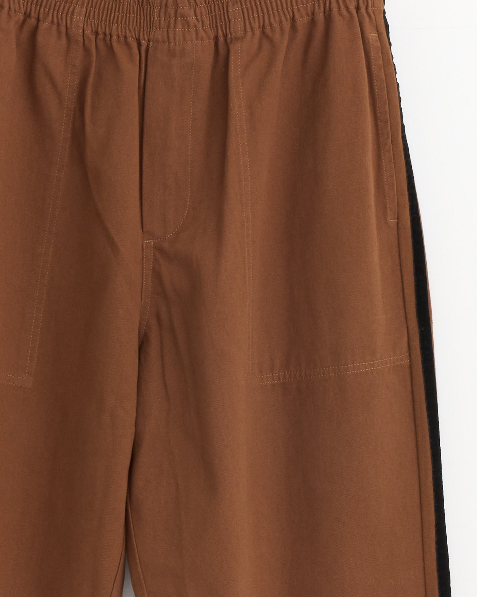 Close-up of Bode's Twill Track Pant in Brown/Black, featuring brown cotton twill, elastic waistband, visible stitching, side pockets, and a black velvet ribbon stripe along the right seam. Set against a plain, light-colored background.