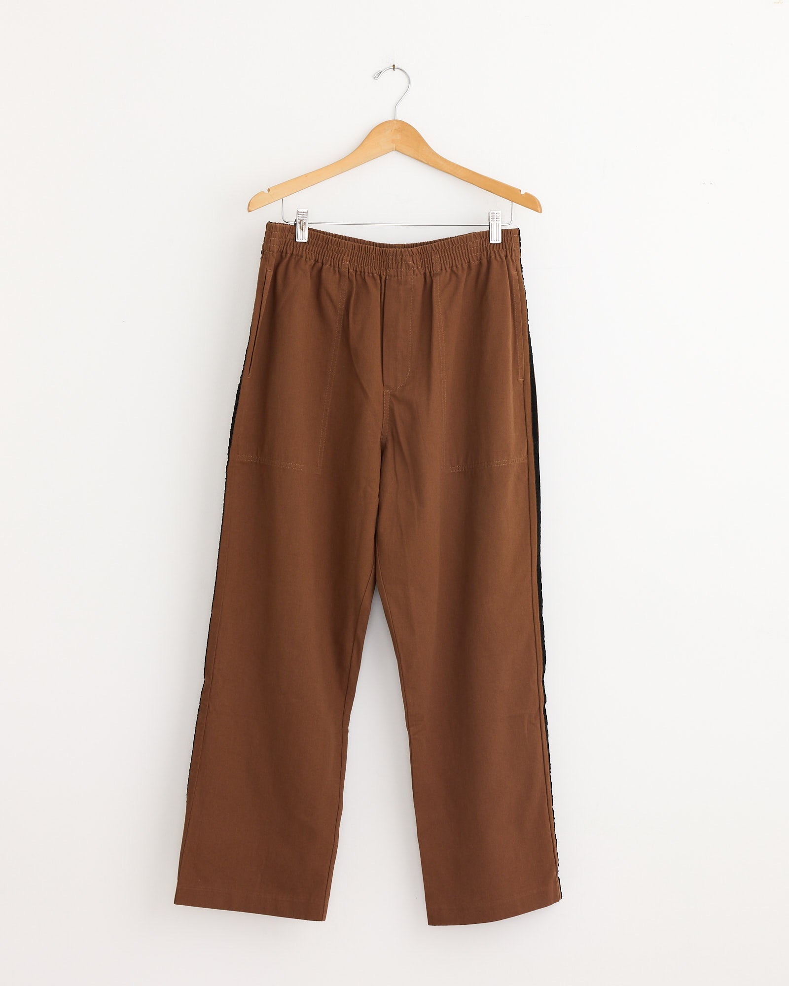 The Bode Twill Track Pant in Brown/Black hangs on a wooden hanger against a white background, showcasing its straight-leg cut, elastic waistband, and visible seam detailing for a casual and comfortable look.