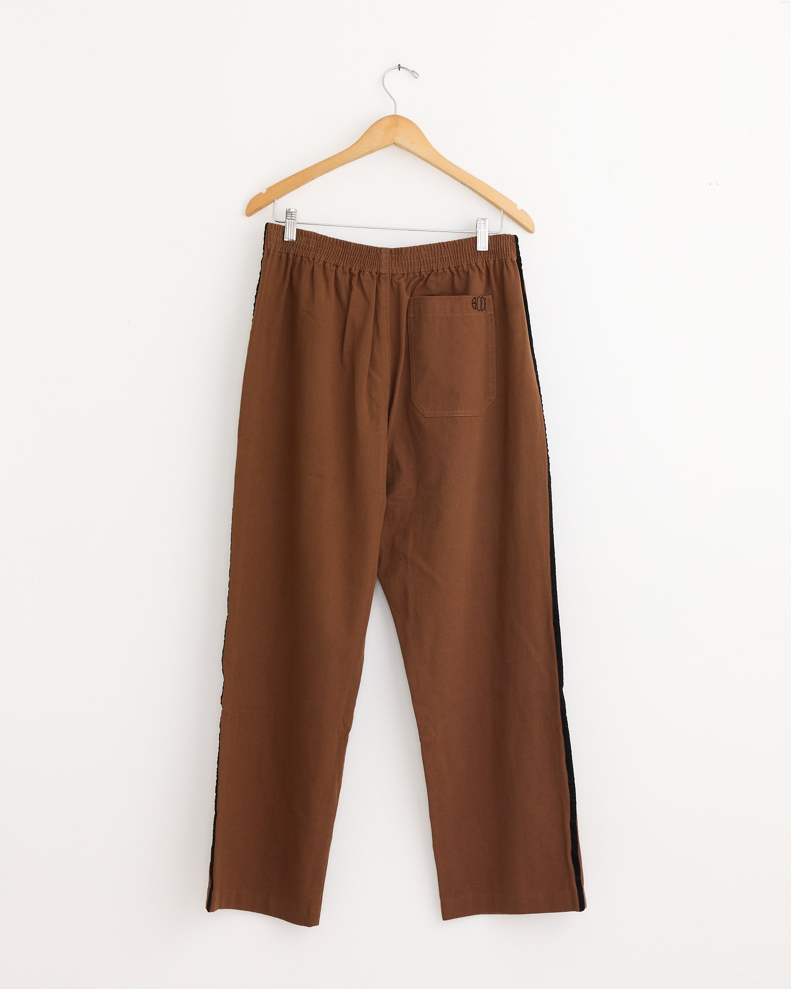 The Bode Twill Track Pant in Brown/Black, featuring an elastic waistband and single back pocket, hangs on a wooden hanger against a plain white background.