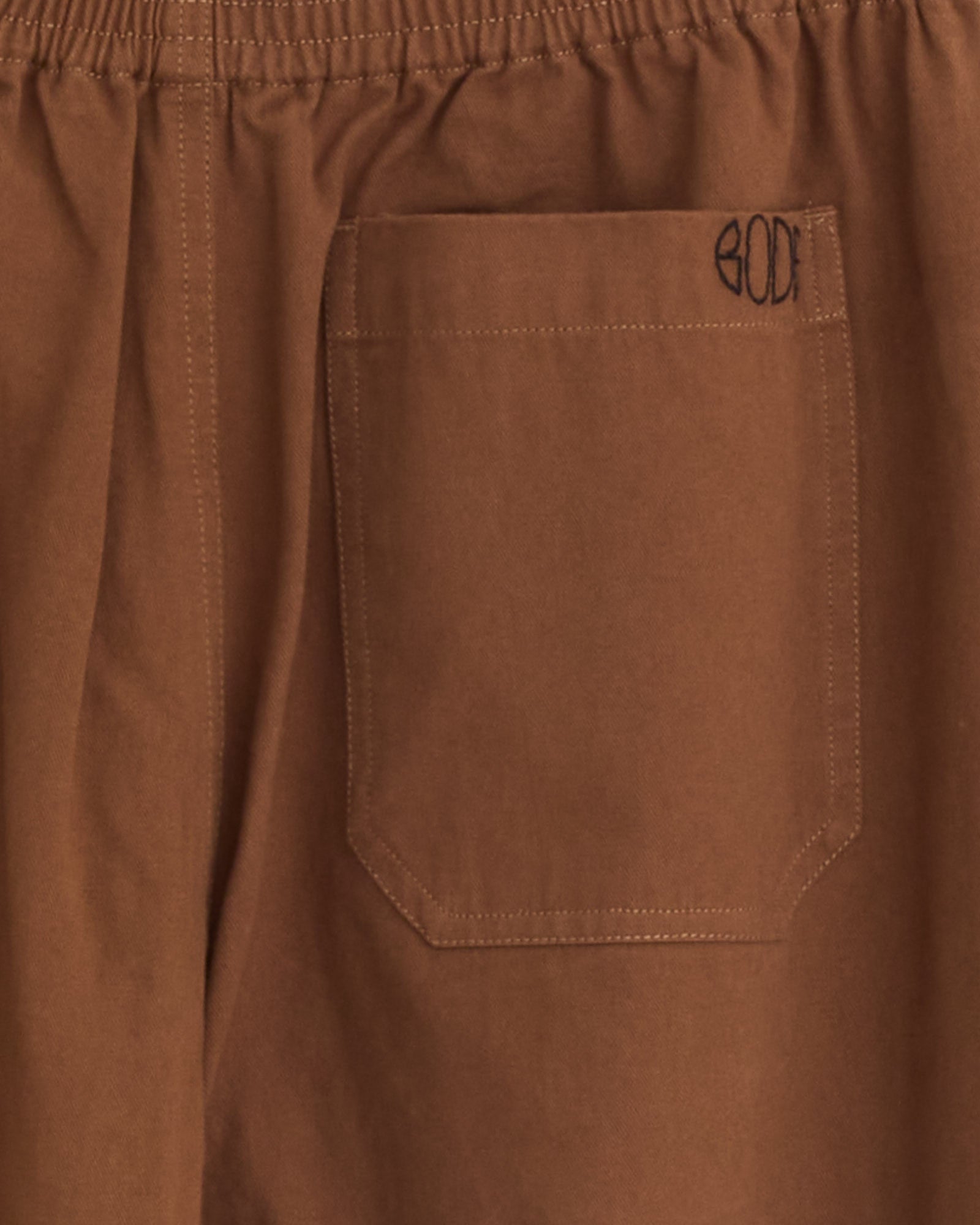 Close-up of the Bode Twill Track Pant in Brown/Black, featuring cotton twill fabric, an elastic waistband, and a rectangular patch pocket with "BODE" stitched in black thread above the pocket.