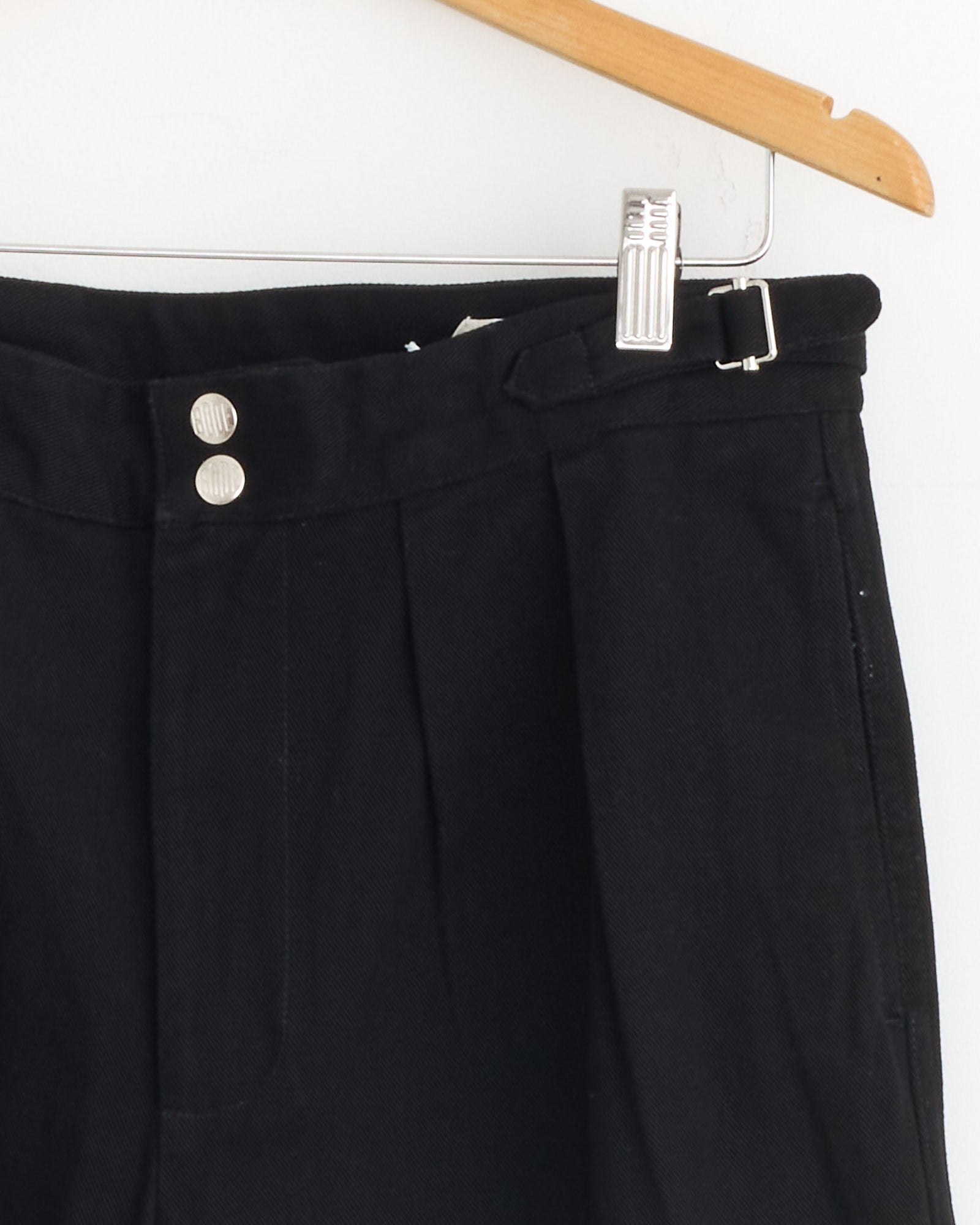 Close-up of the upper part of Bode’s Wide Leg Snap Trouser in Black, heavyweight 100% cotton twill with two silver buttons, a belt loop with buckle, pleated detailing, and wide-leg silhouette hanging on a wooden hanger.