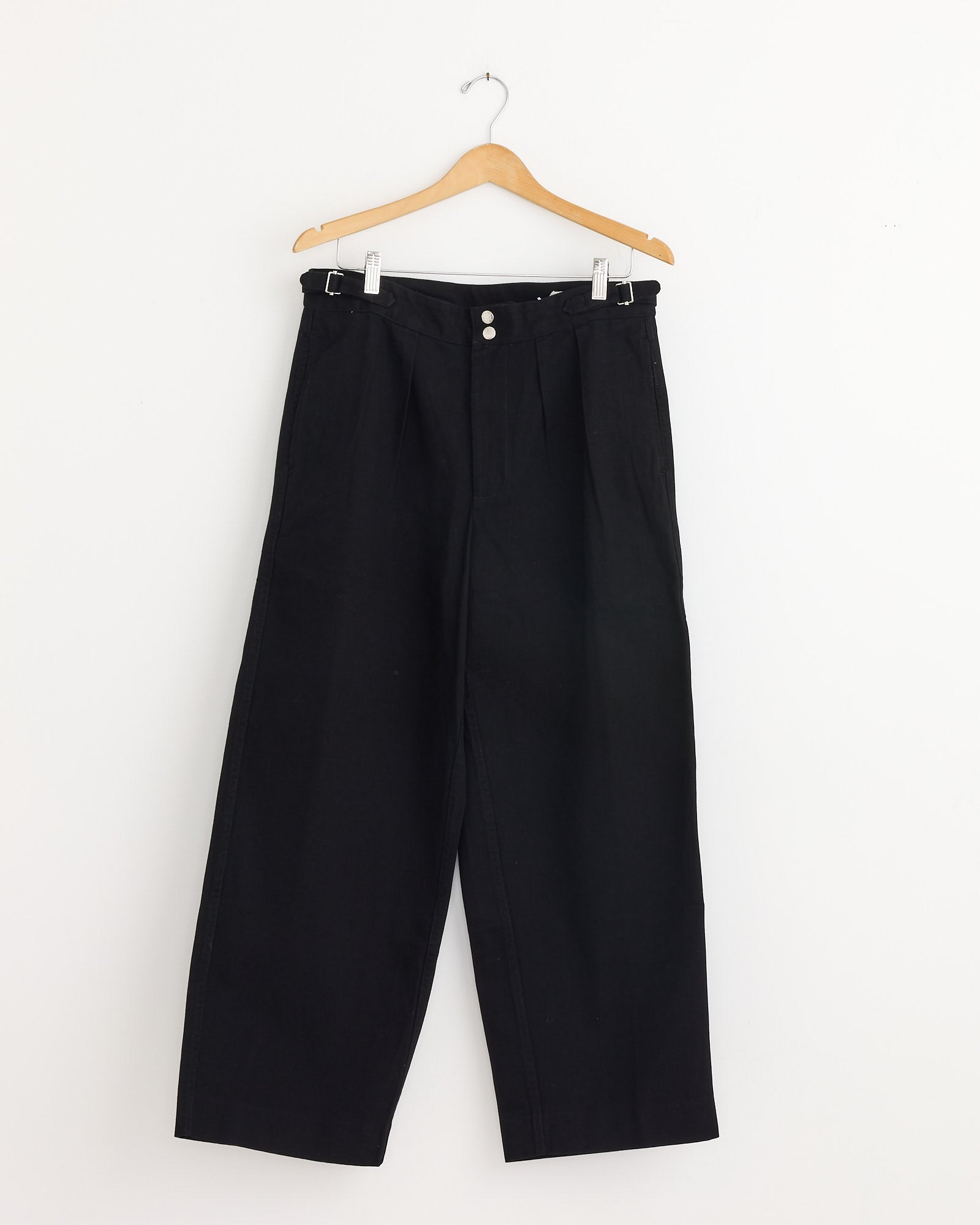 The Bode Wide Leg Snap Trouser in Black, a high-waisted heavyweight twill style with two buttons and belt loops, hangs on a wooden hanger against a plain white background.