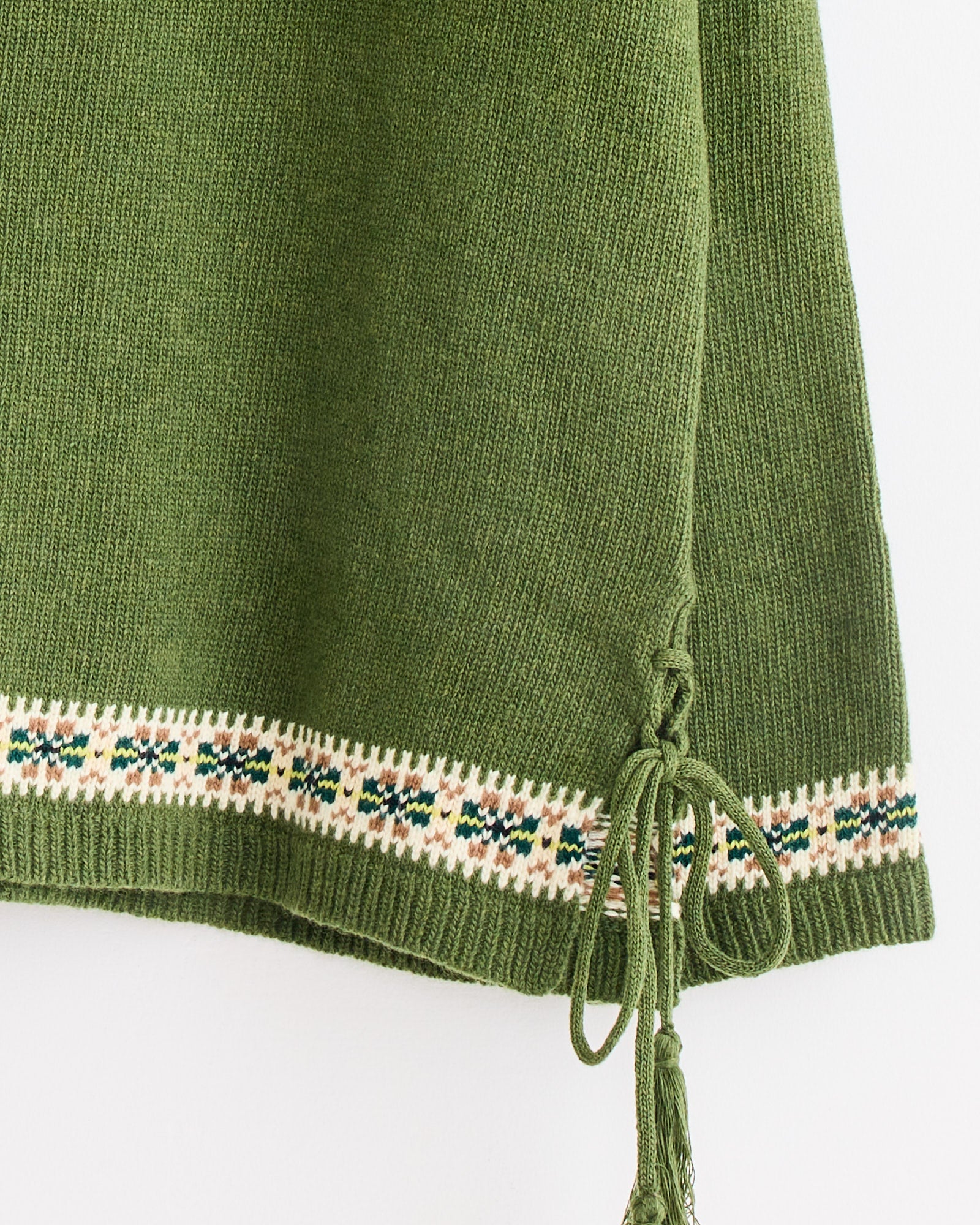 Close-up of the Moritz Sweater in Green Multi by Bode, featuring green Fair Isle knit with a cream, navy, and green border, plus a tie detail with a small bow and tassel at the lower right edge.