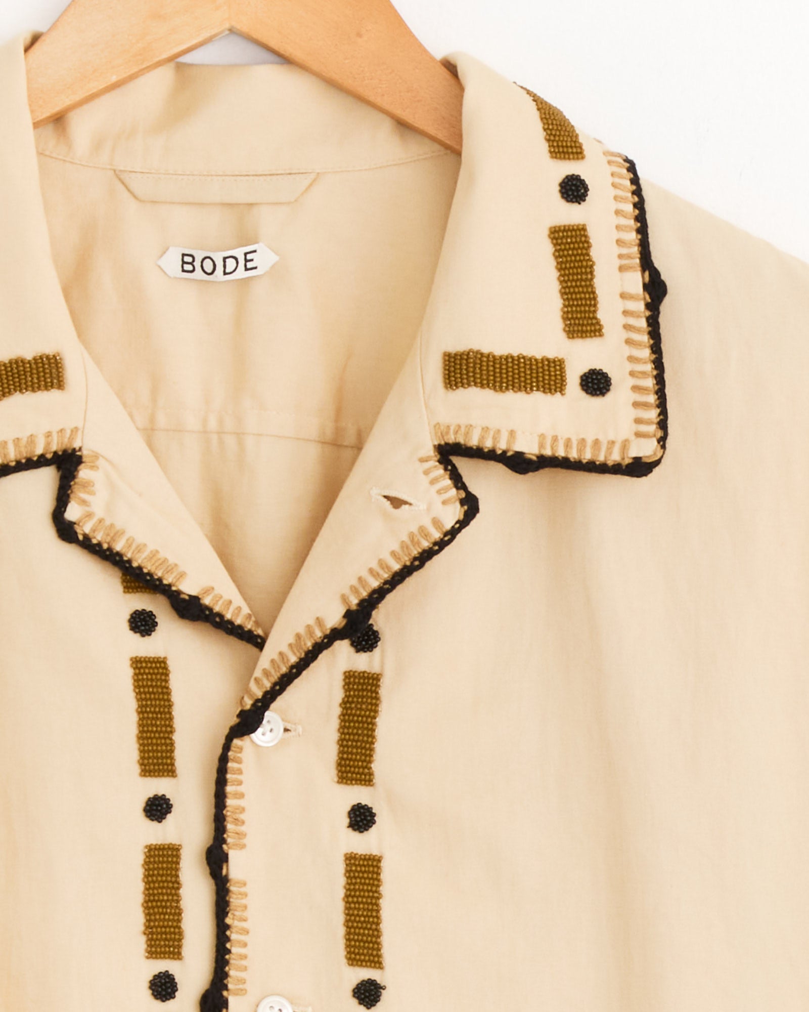 The Beaded Ryegrass Shirt in Brown by Bode hangs on a wooden hanger, showcasing handmade olive-green geometric beadwork and black stitched accents on the collar and button placket. The inner label reads "Bode.