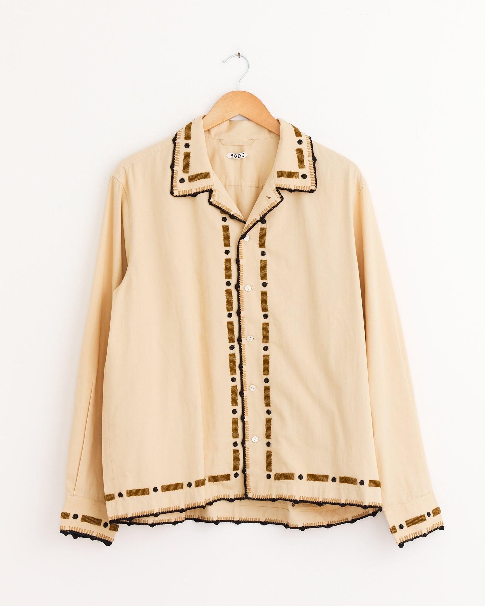 The Bode Beaded Ryegrass Shirt in Brown, featuring long sleeves, brown geometric trim, and handmade beadwork on the collar, placket, cuffs, and hem, hangs on a wooden hanger against a white wall.