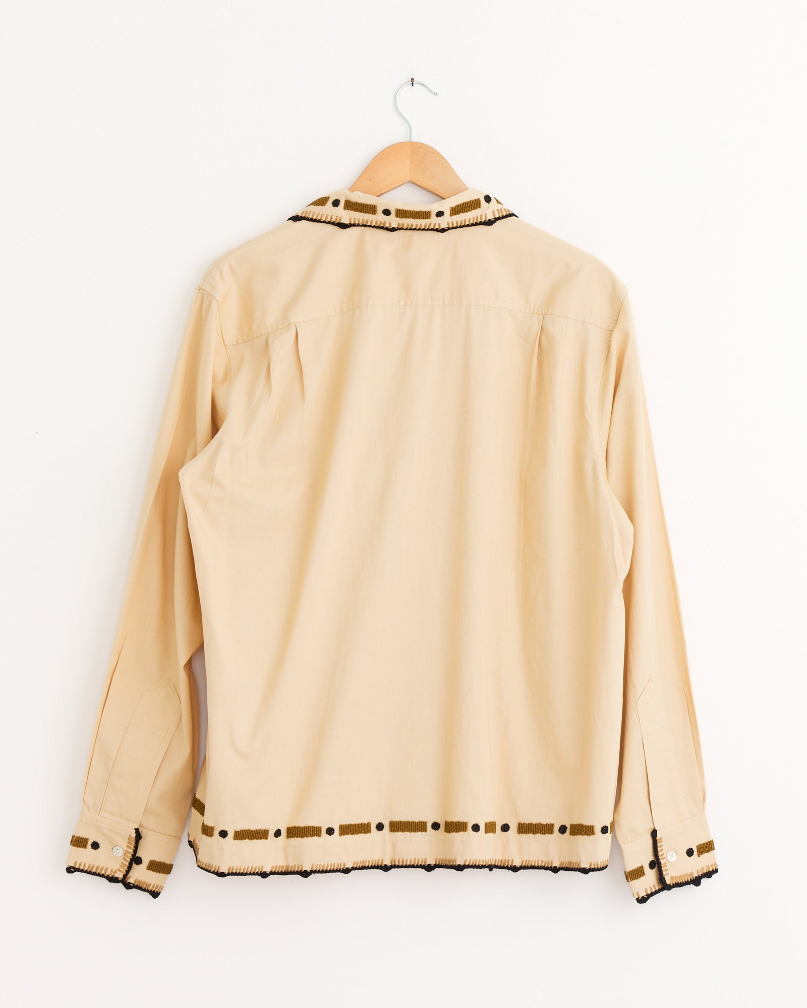 The Bode Beaded Ryegrass Shirt in Brown features black trim and handmade beadwork on the collar, cuffs, and hem. Displayed backwards on a wooden hanger against a plain white wall.