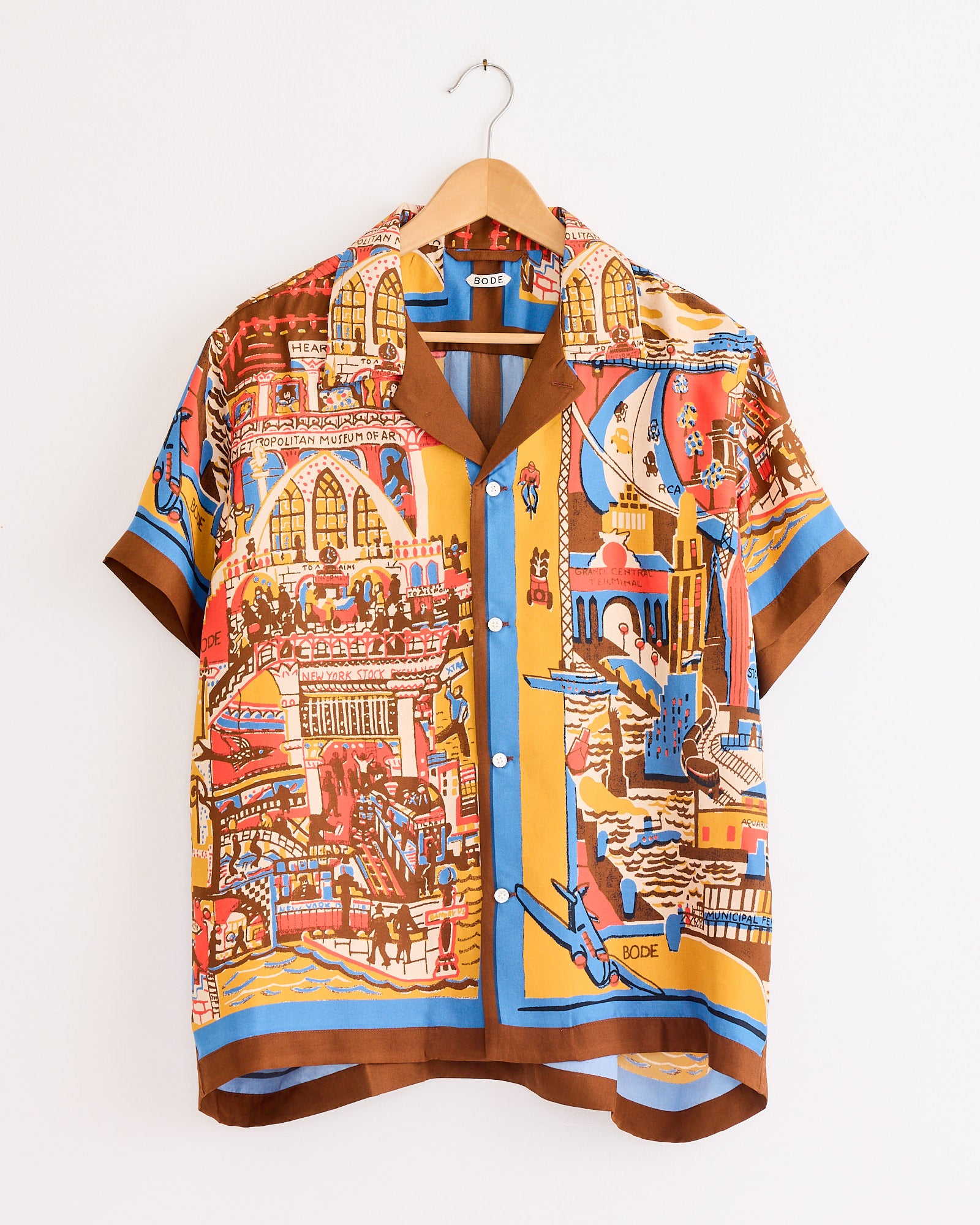 The Bode Manhattan Island Shirt in Multi is a short-sleeved silk button-up with a colorful cityscape print, featuring buildings, people, and boats in red, yellow, blue, and brown. Shown on a wooden hanger against a white wall.