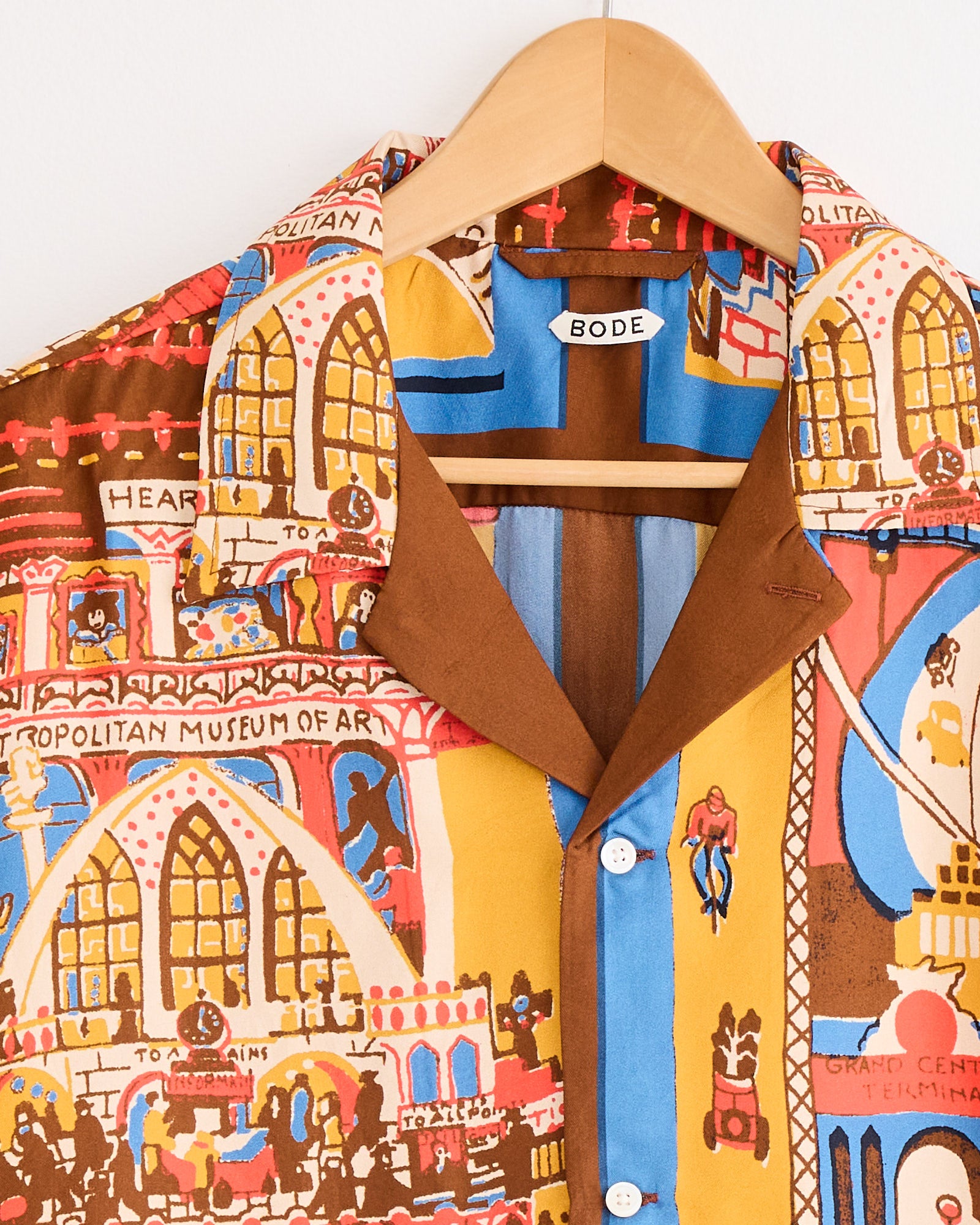 The Bode Manhattan Island Shirt in Multi hangs on a wooden hanger and showcases illustrated buildings, including the Metropolitan Museum of Art, in brown, blue, yellow, red, and cream tones.