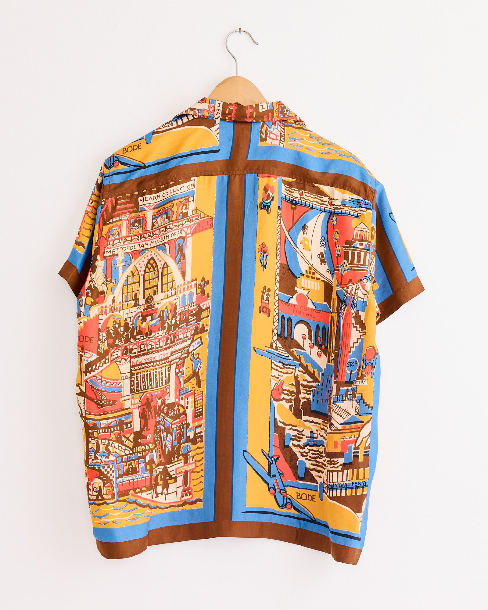 The Bode Manhattan Island Shirt in Multi hangs on a wooden hanger, showcasing bold graphic illustrations of cityscapes and boats in red, yellow, blue, and brown on a white silk background.