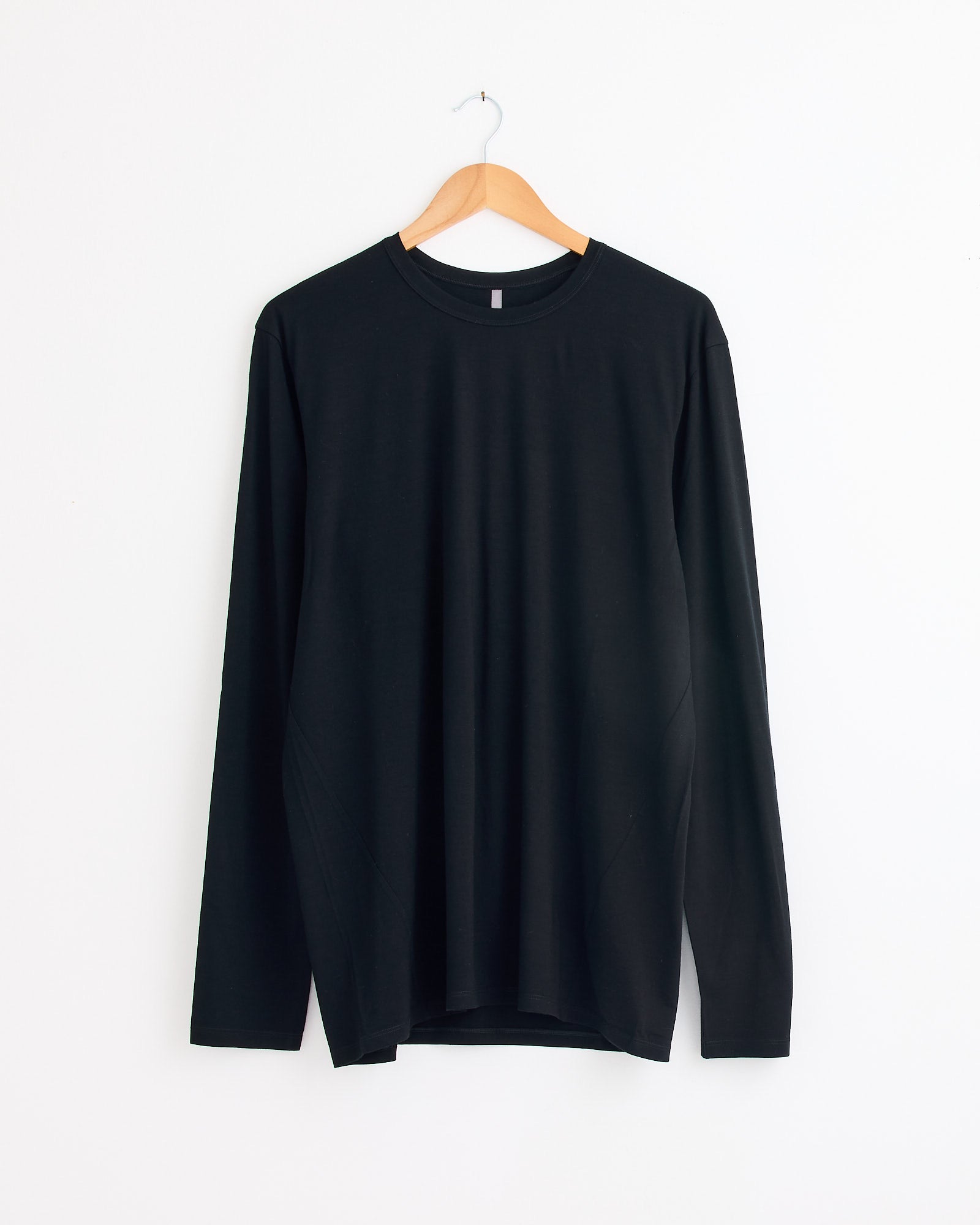 The Veilance Frame Shirt in Black, made from ultrafine Merino wool, hangs on a wooden hanger against a white wall, providing natural odour resistance and temperature regulation.