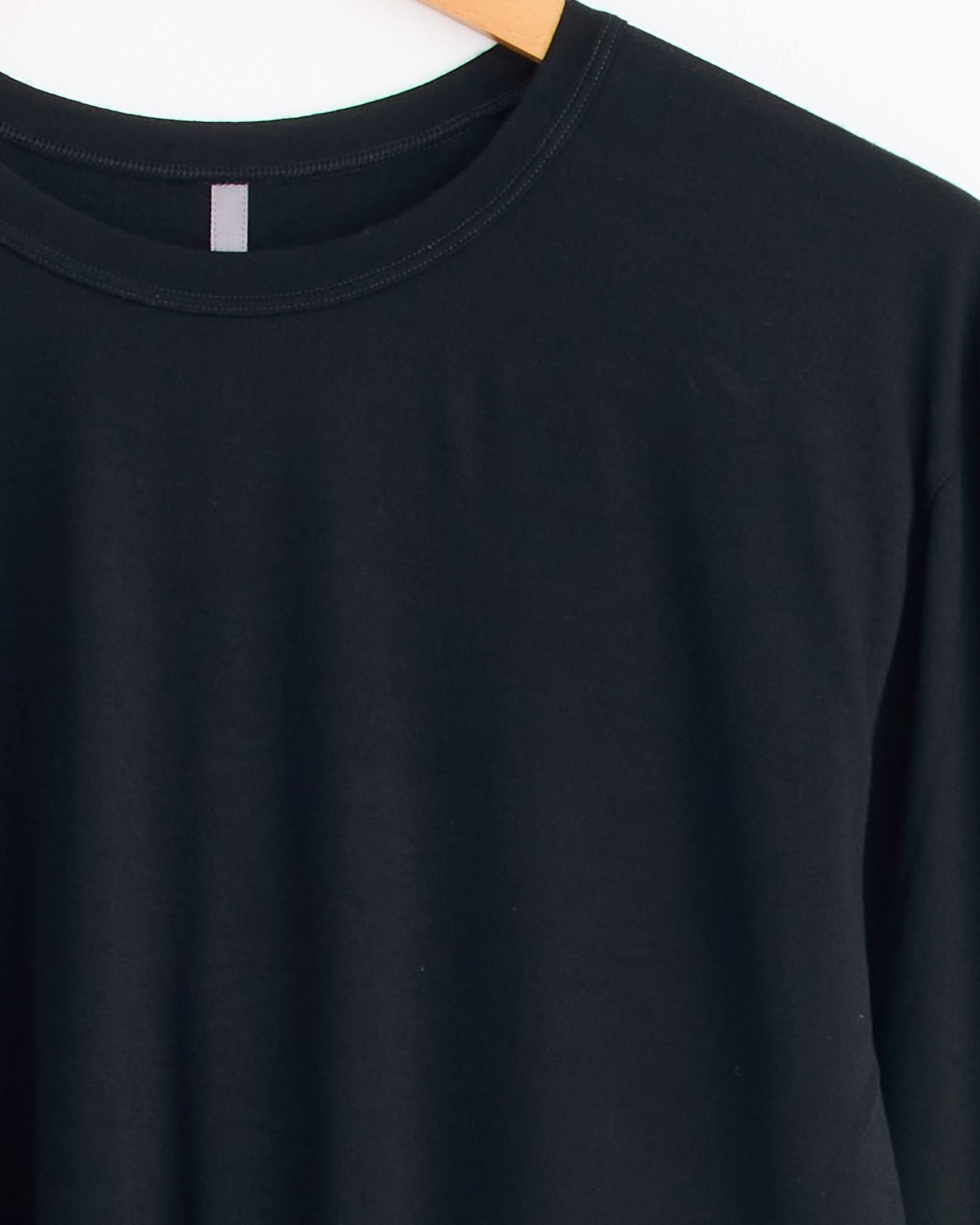 The Veilance Frame Shirt in Black, crafted from ultrafine Merino wool, hangs on a wooden hanger against a white background, highlighting its upper chest, neckline, and part of the left sleeve for exceptional comfort and temperature control.