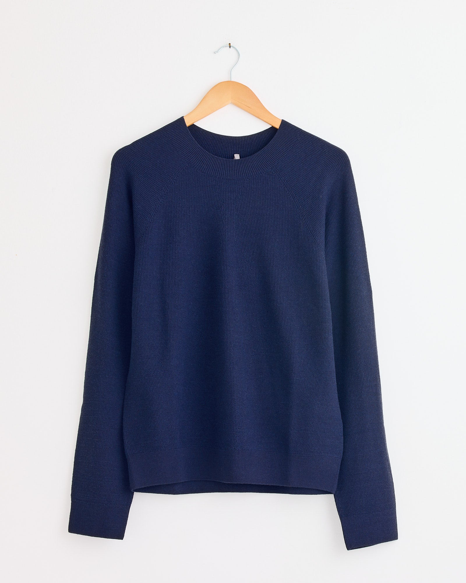The Conic Wool Midlayer in Passport by Veilance, a dark blue long-sleeve knit sweater, hangs on a wooden hanger against a plain white wall. For sizing questions, reach out via our online chat.