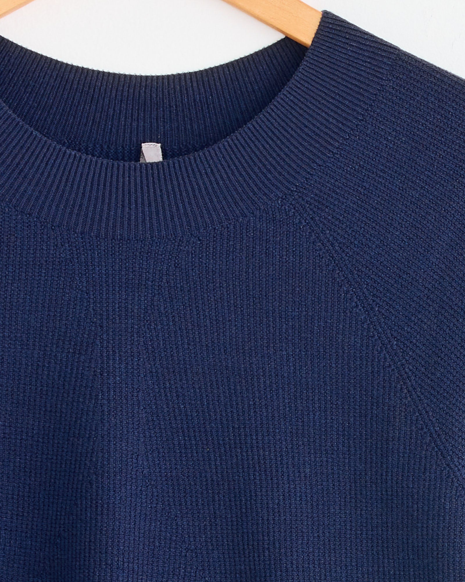 Close-up of the Veilance Conic Wool Midlayer in Passport, featuring a ribbed crew neckline, hanging on a light wooden hanger against a white background. For fit or sizing questions, connect with us via online chat.