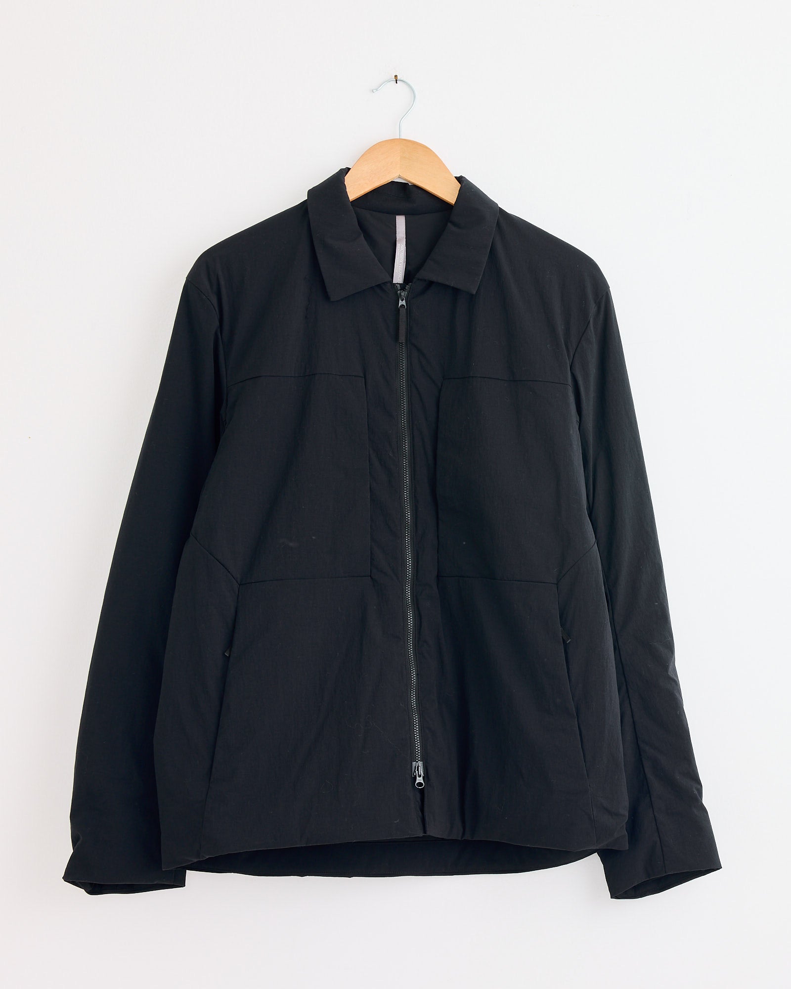 The Veilance Mionn Insulated Overshirt in Black, featuring a collar and zip-up front, hangs on a wooden hanger against a plain white wall. For sizing or fit questions, contact us via online chat for quick help.