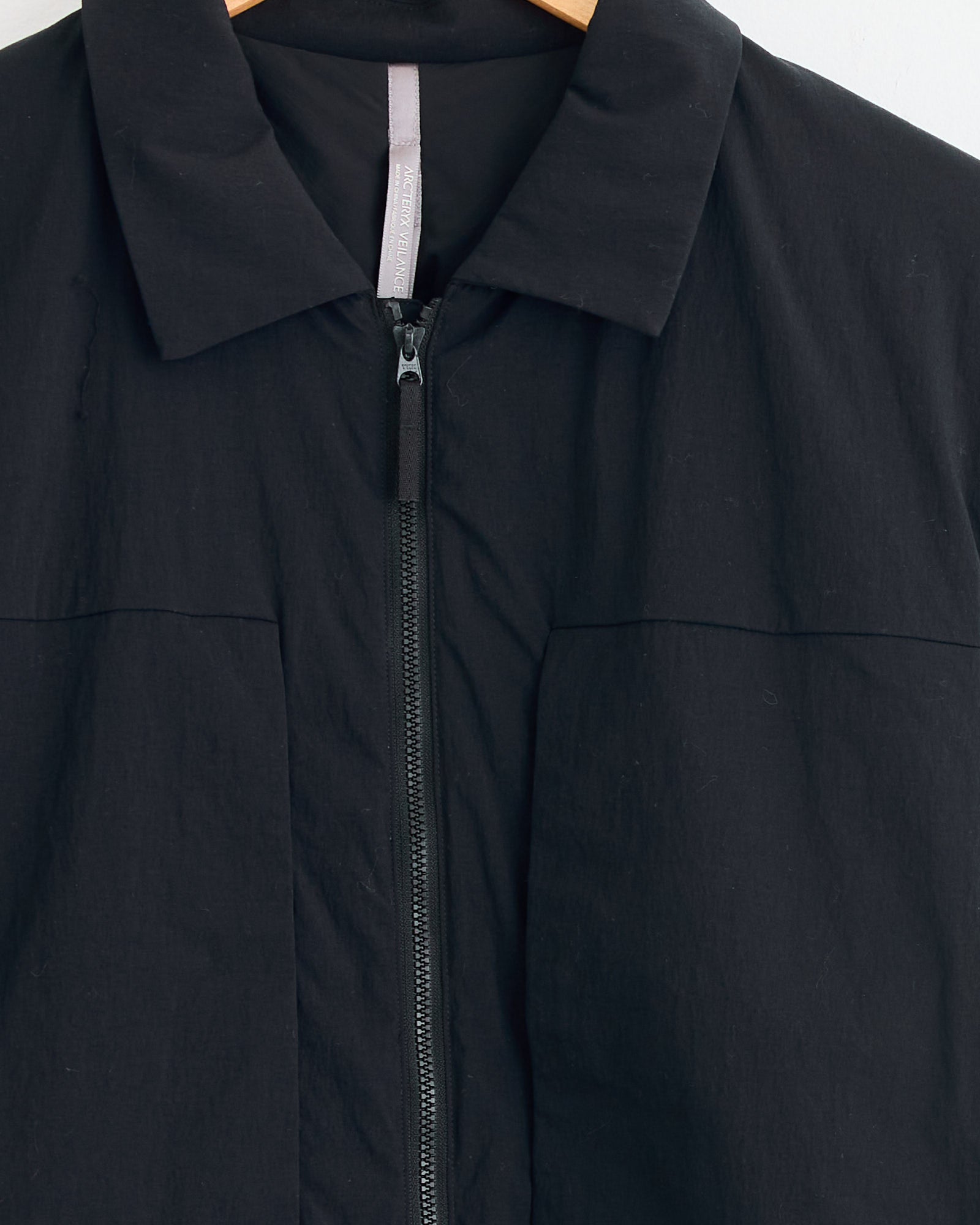 Close-up of the Veilance Mionn Insulated Overshirt in Black, featuring a collar and front zipper, displayed on a wooden hanger with a light tag inside. Need sizing help? Chat with us online for expert assistance.