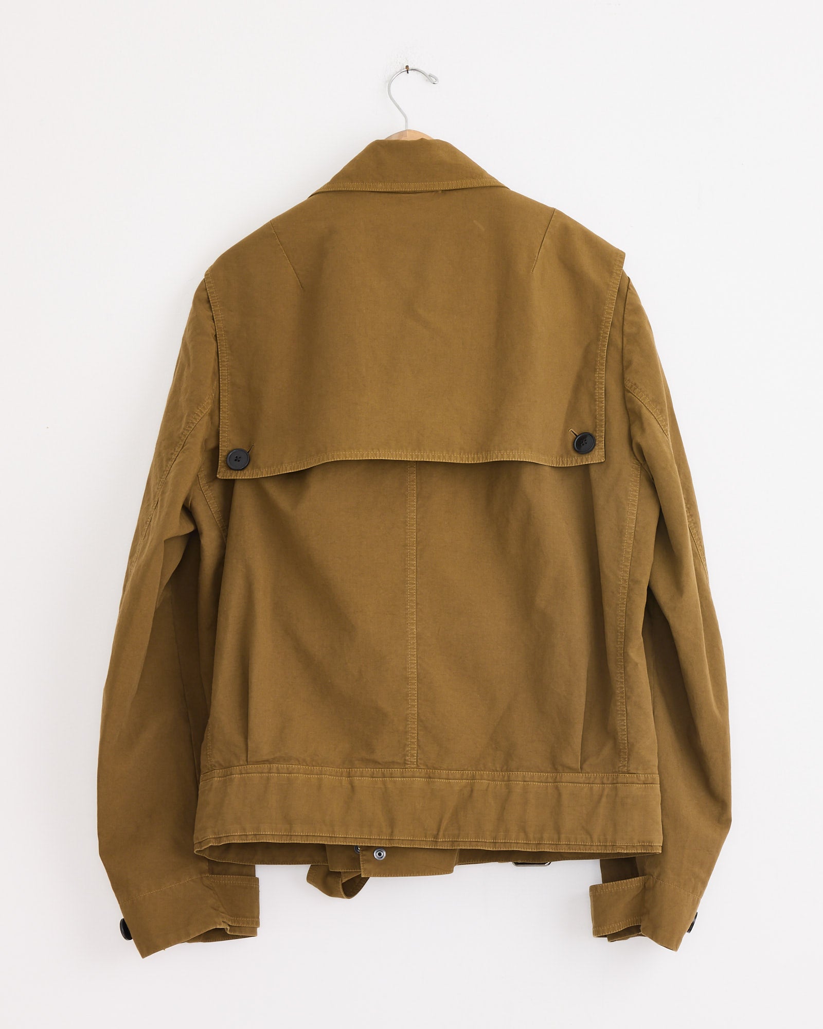 A Dries Van Noten Belted Biker Jacket in olive hangs on a hook against a white wall, displayed with the back facing forward. The jacket features a wide collar, button details, and long sleeves.
