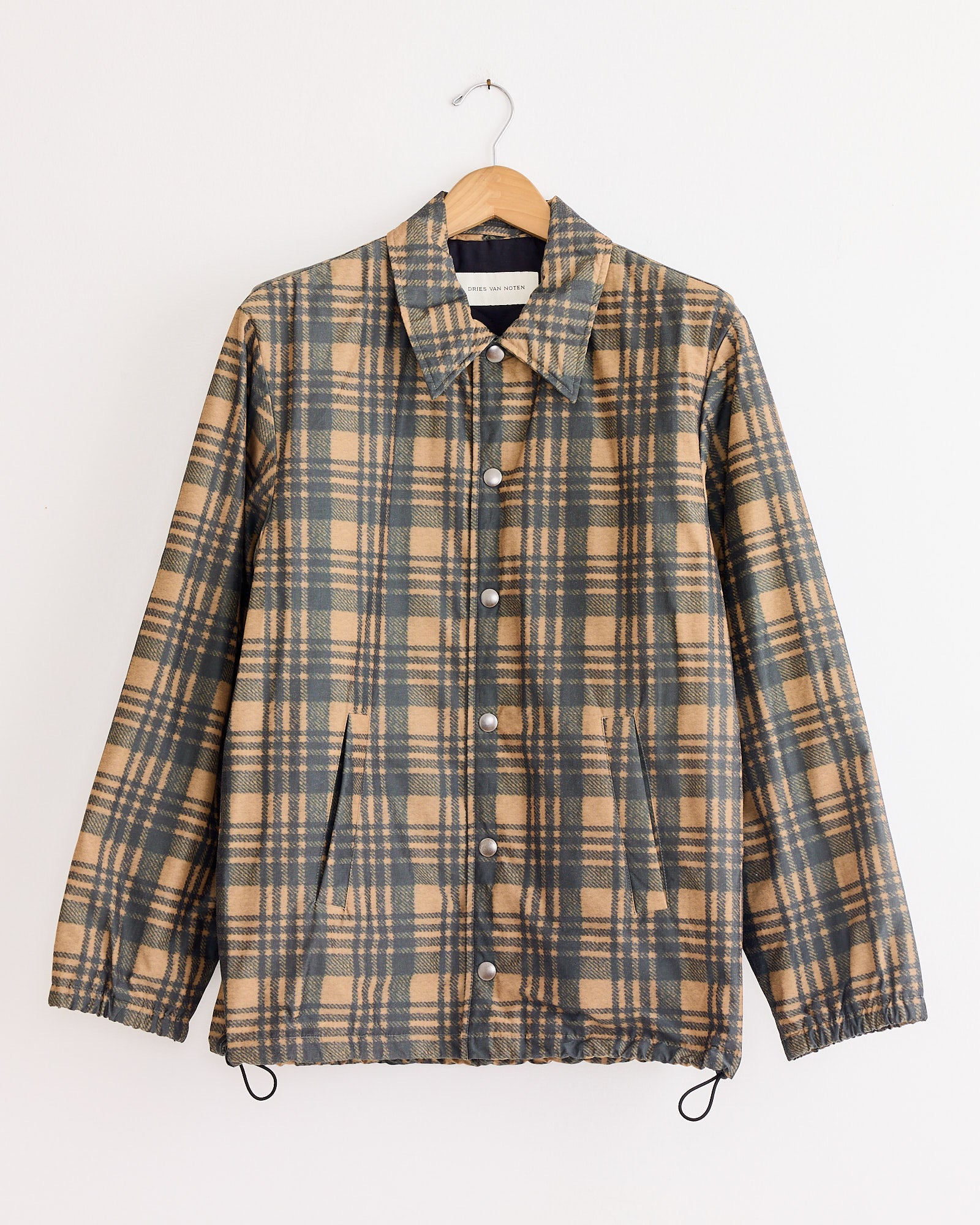 The Dries Van Noten Vorries 2026 Jacket in Tan, a beige and dark gray plaid polyester-cotton piece with a collar, button-down front, lining, two slanted pockets, and elastic cuffs hangs on a wooden hanger against a white wall.