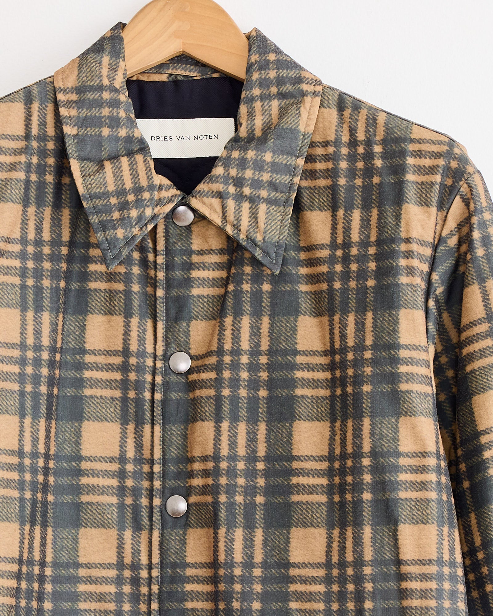 The Dries Van Noten Vorries 2026 Jacket in tan, featuring a beige and dark green plaid polyester-cotton blend with silver snap buttons, hangs on a wooden hanger. The label inside the collar reads Dries Van Noten.