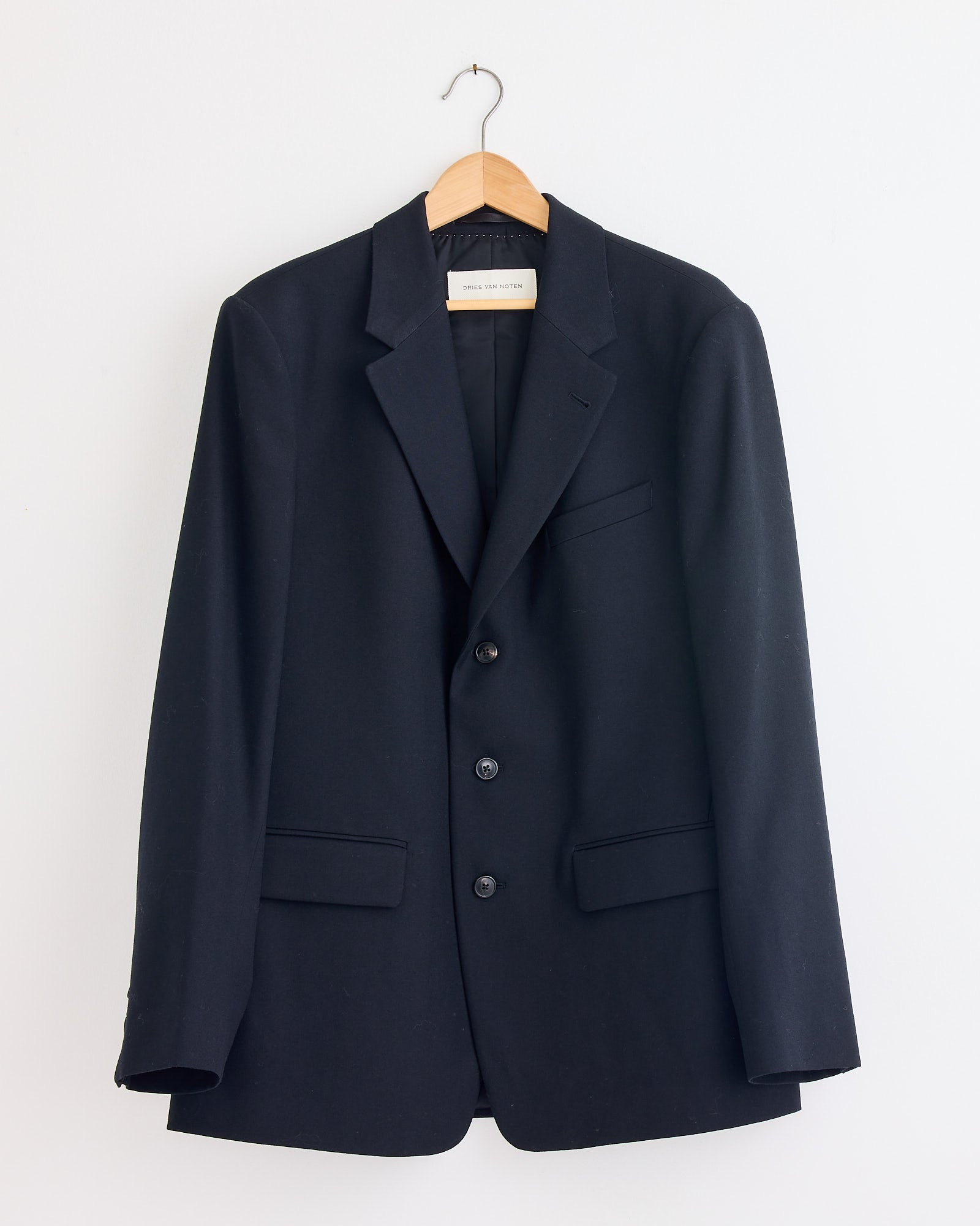 The Dries Van Noten Kiel 2246 Suit in Black, a classic wool blazer with three buttons and two front pockets, hangs on a wooden hanger against a white wall.